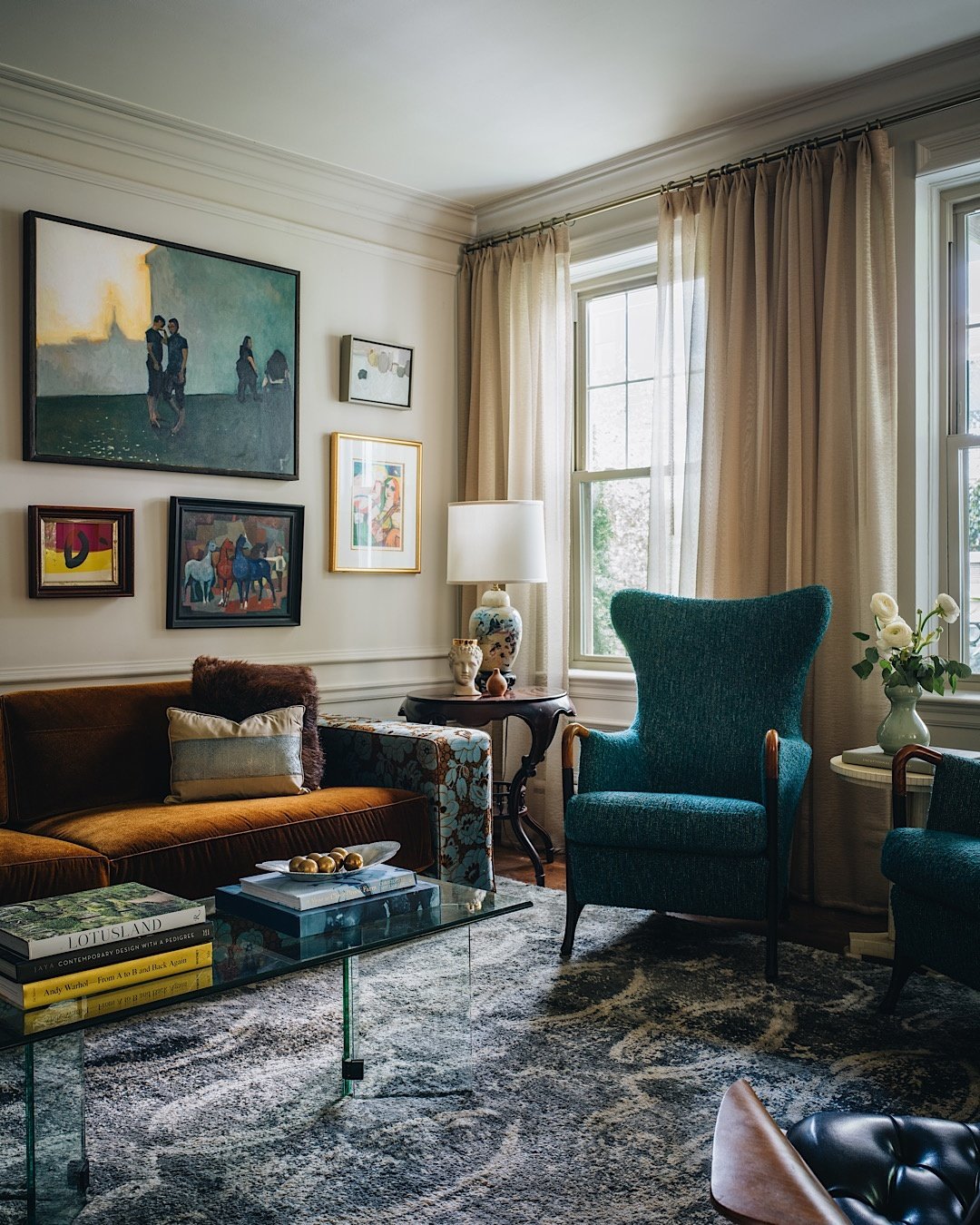 Eclectic salon-style gallery wall with various sized framed paintings hung above a brown velvet sofa and next to a teal wingback chair.