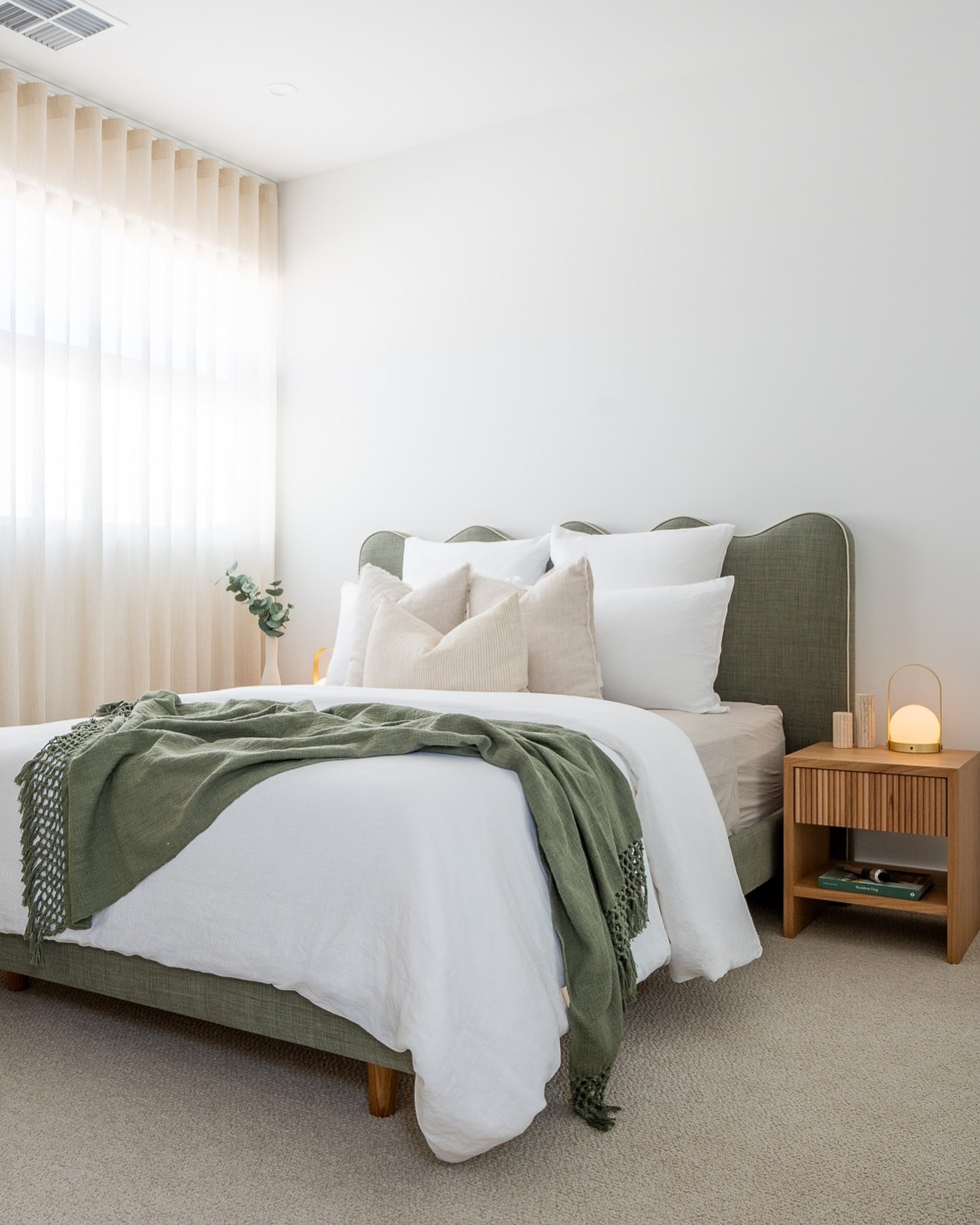 Bedroom with a scalloped sage green upholstered bed, white and beige layered bedding, a matching green knit throw, and a slatted wooden nightstand.