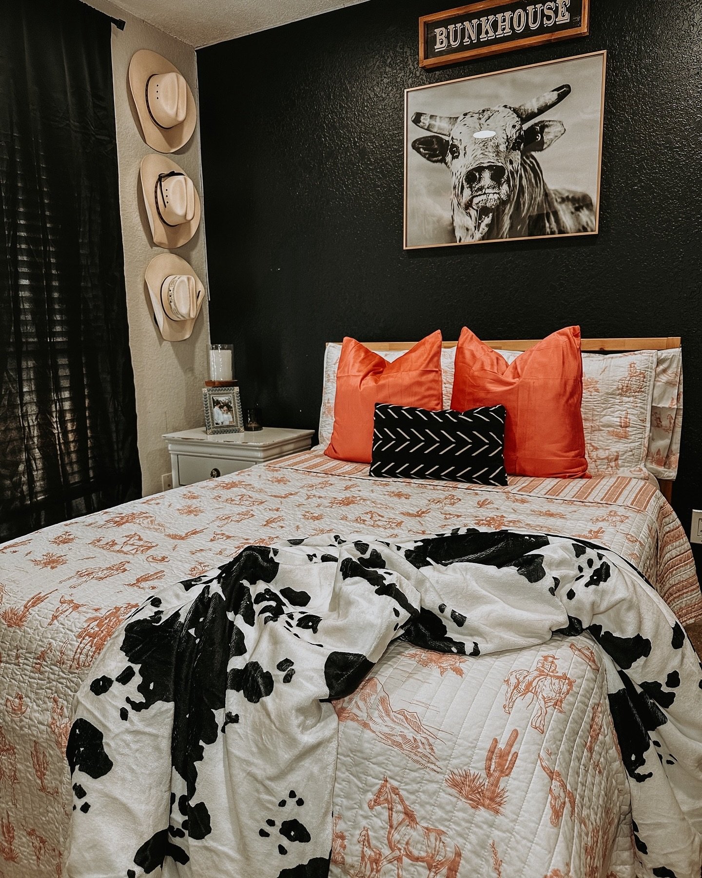 A Western bedroom with a textured black accent wall, a framed bull photo, bright orange throw pillows, and cow print bedding.