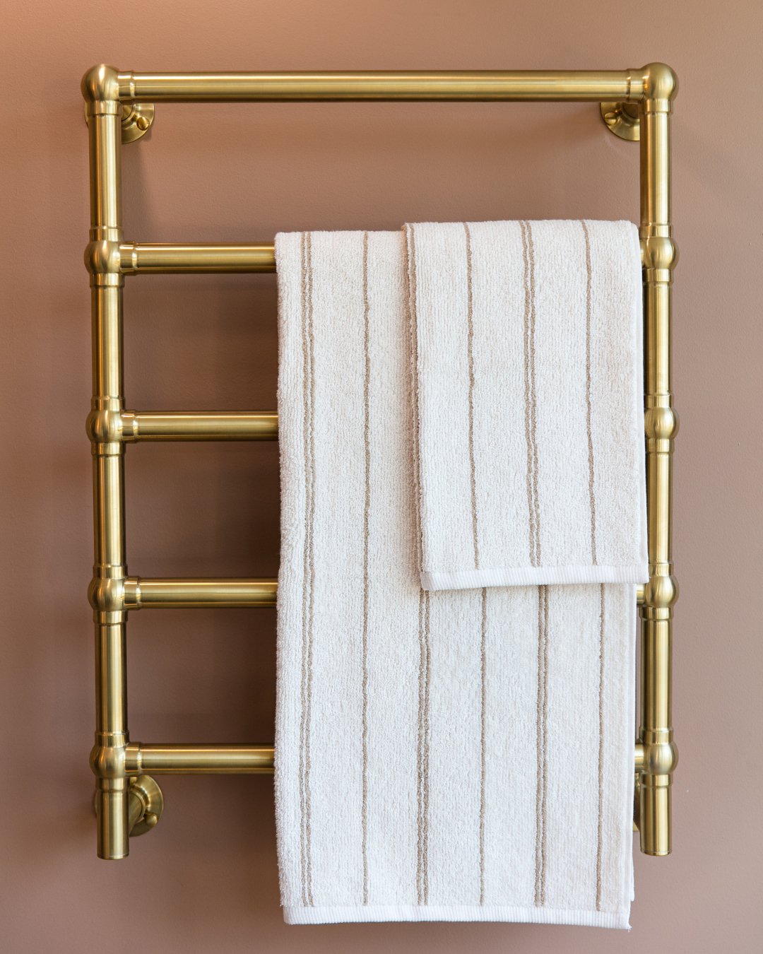 Close-up of a traditional style gold or brass heated towel radiator with visible plumbing joints, holding a white striped bath towel against a warm beige wall.
