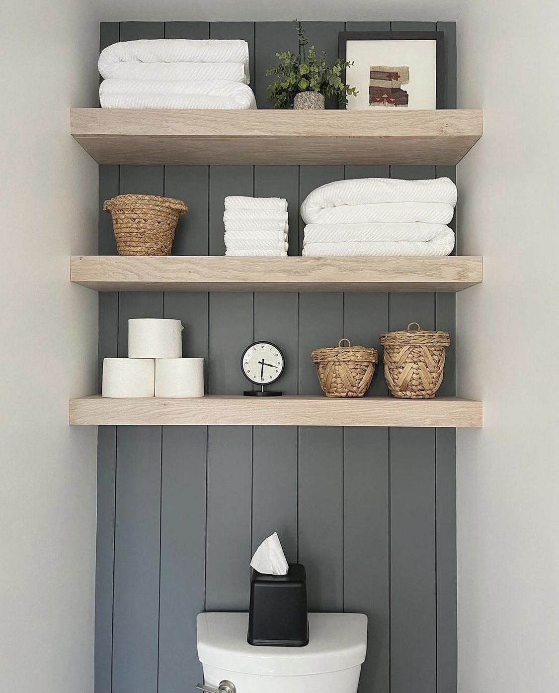 Three light wood floating shelves installed over a dark gray vertical shiplap accent panel, styled with baskets and towels.