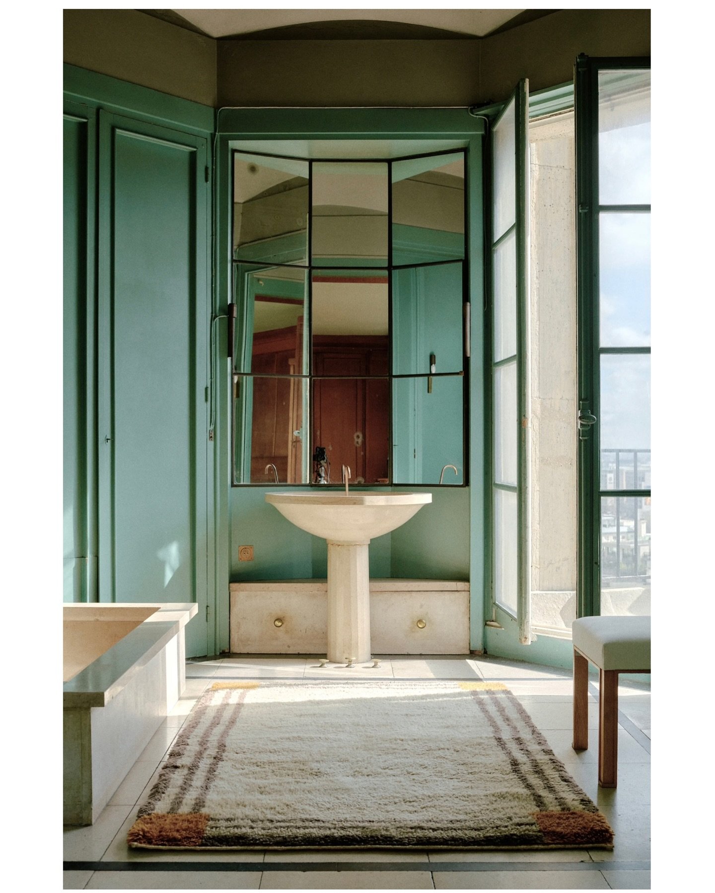 A striking bathroom with mint green walls, a black-framed segmented mirror, a pedestal sink, and a rectangular off-white, shaggy rug with two thin brown border stripes.