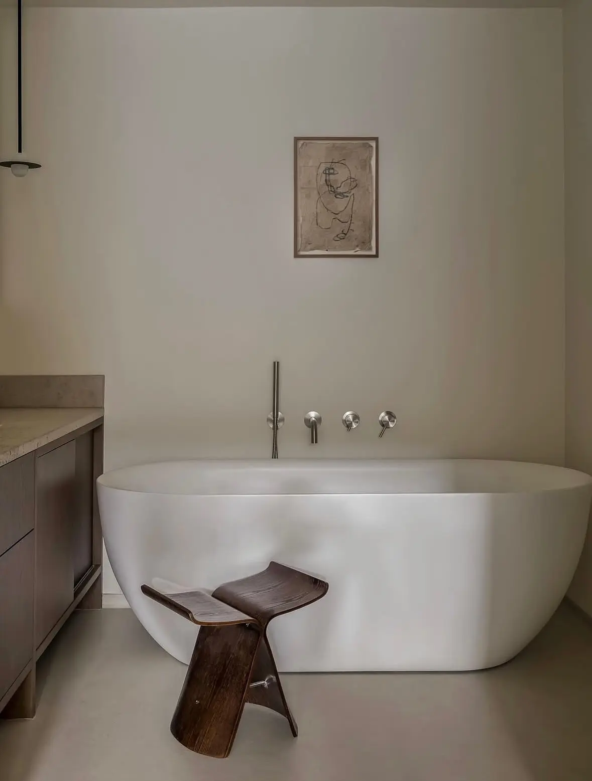 A neutral, contemporary bathroom with a white freestanding tub, featuring a small framed piece of simple, single-line figurative art.