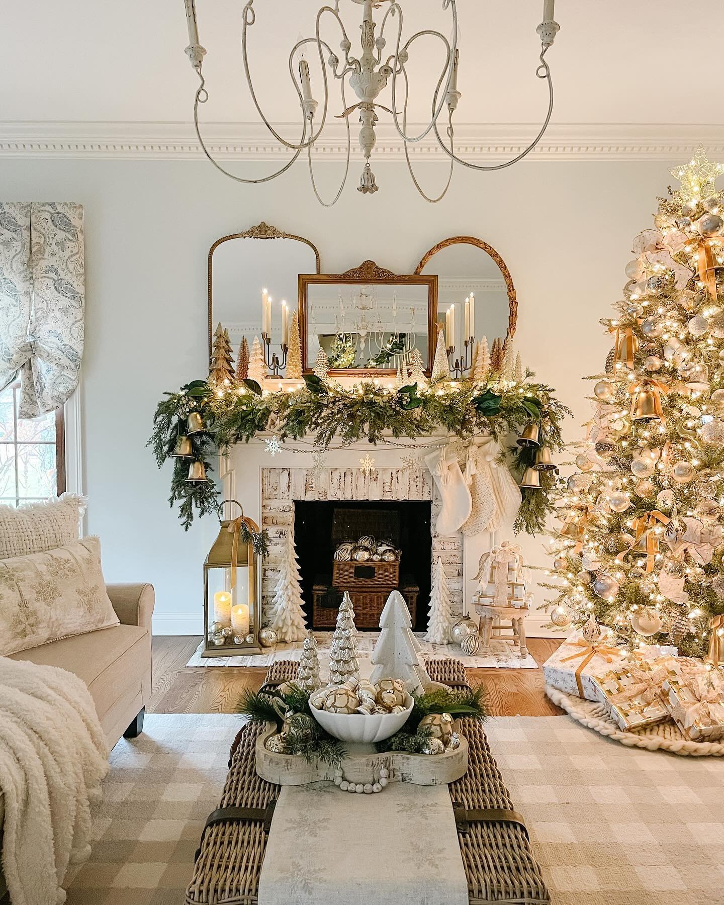 White brick fireplace with three gold mirrors above, decorated with a thick pine garland, white knit stockings, and large hanging gold bells.