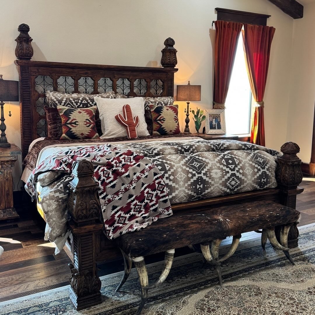 A grand Western bedroom featuring an intricately carved dark wood four-poster bed, layered Southwestern bedding, and a cowhide bench with real horns for legs.