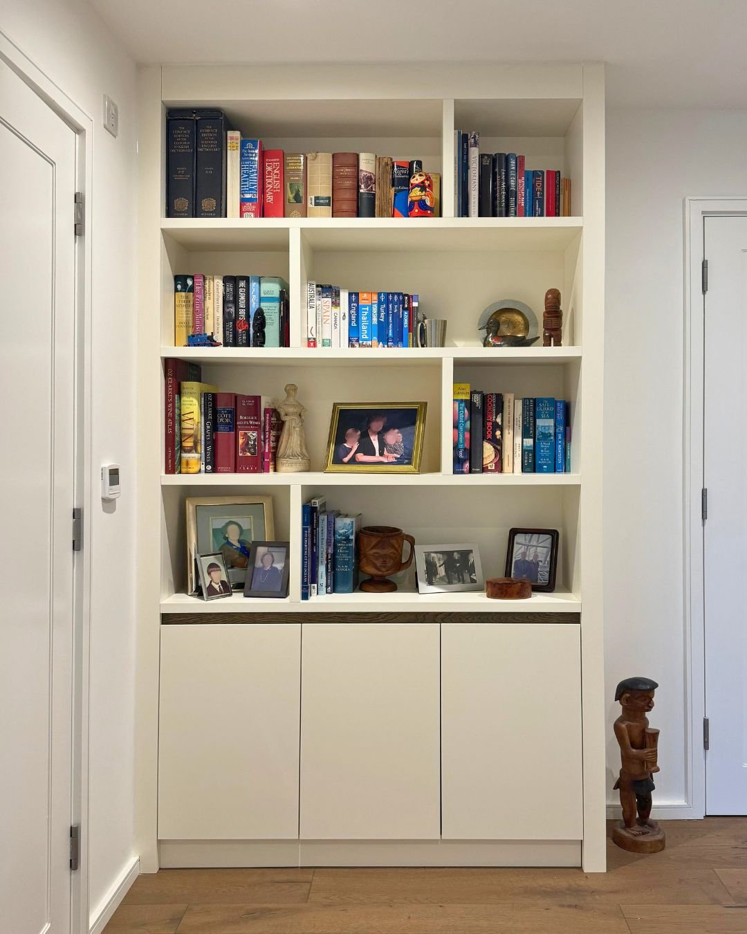 A tall, narrow white built-in storage unit featuring open shelves with varied cubby sizes above a base of three closed cabinets, filled with books and framed photos.