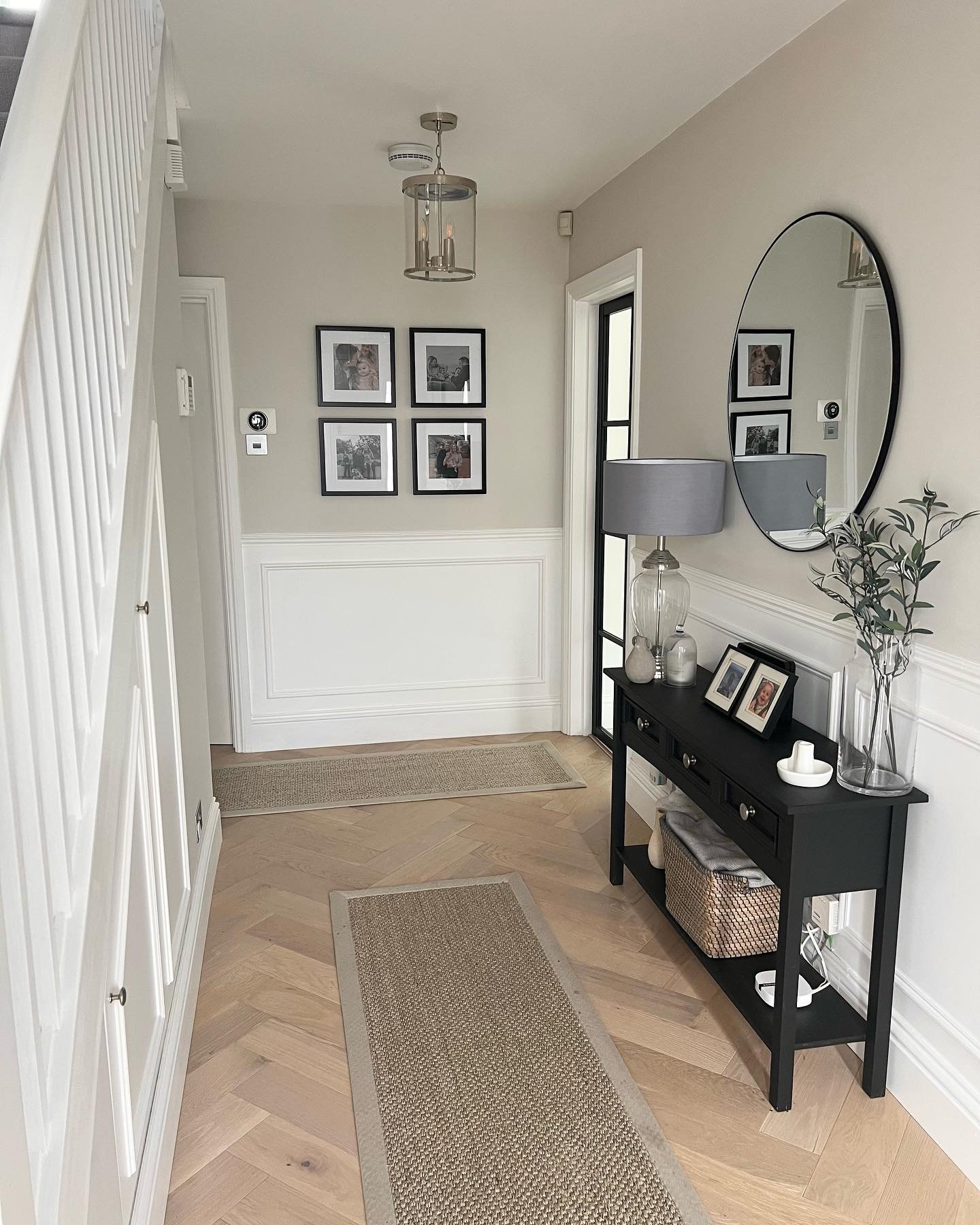 A bright hallway with herringbone wood floor, a black console table, a large round mirror, and white wainscoting on a light taupe wall.