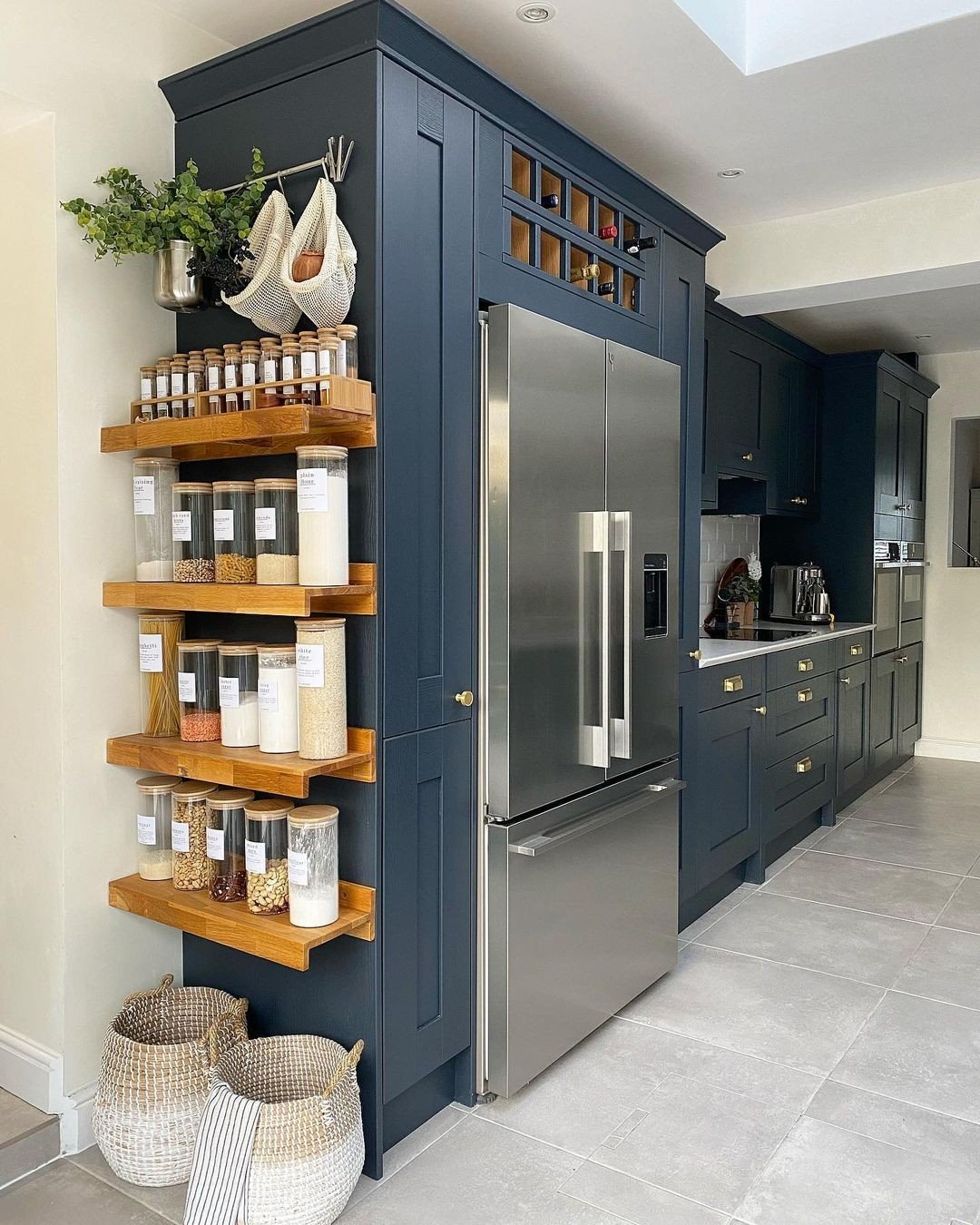 Dark blue kitchen cabinets with thick, natural wood floating shelves on the side, organizing clear jars of dry goods and spices.