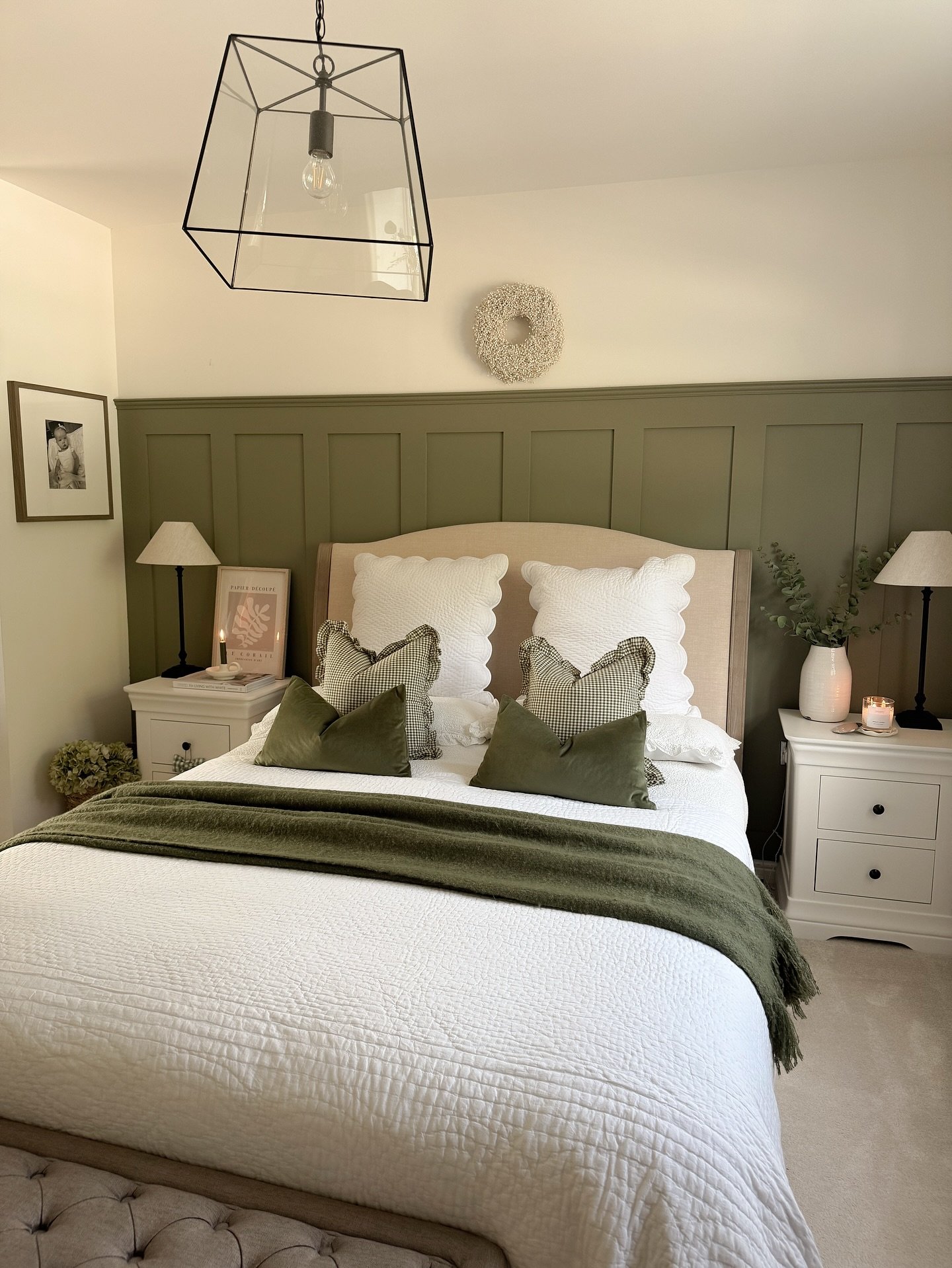A bedroom with a neutral upholstered bed, a board and batten wall painted deep olive green, and white bedside tables.