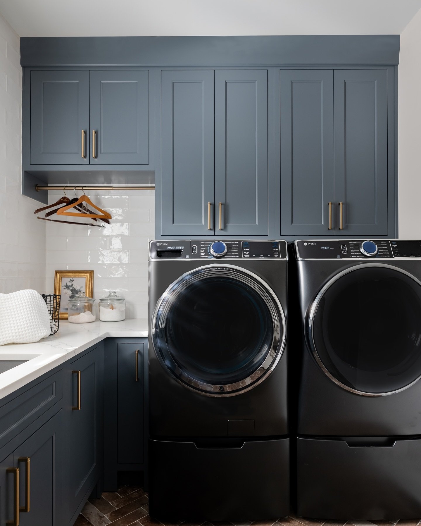 A small, modern laundry room featuring deep navy or slate blue Shaker-style cabinets, dark charcoal appliances on pedestals, white marble-look counter, and a subway tile backsplash.