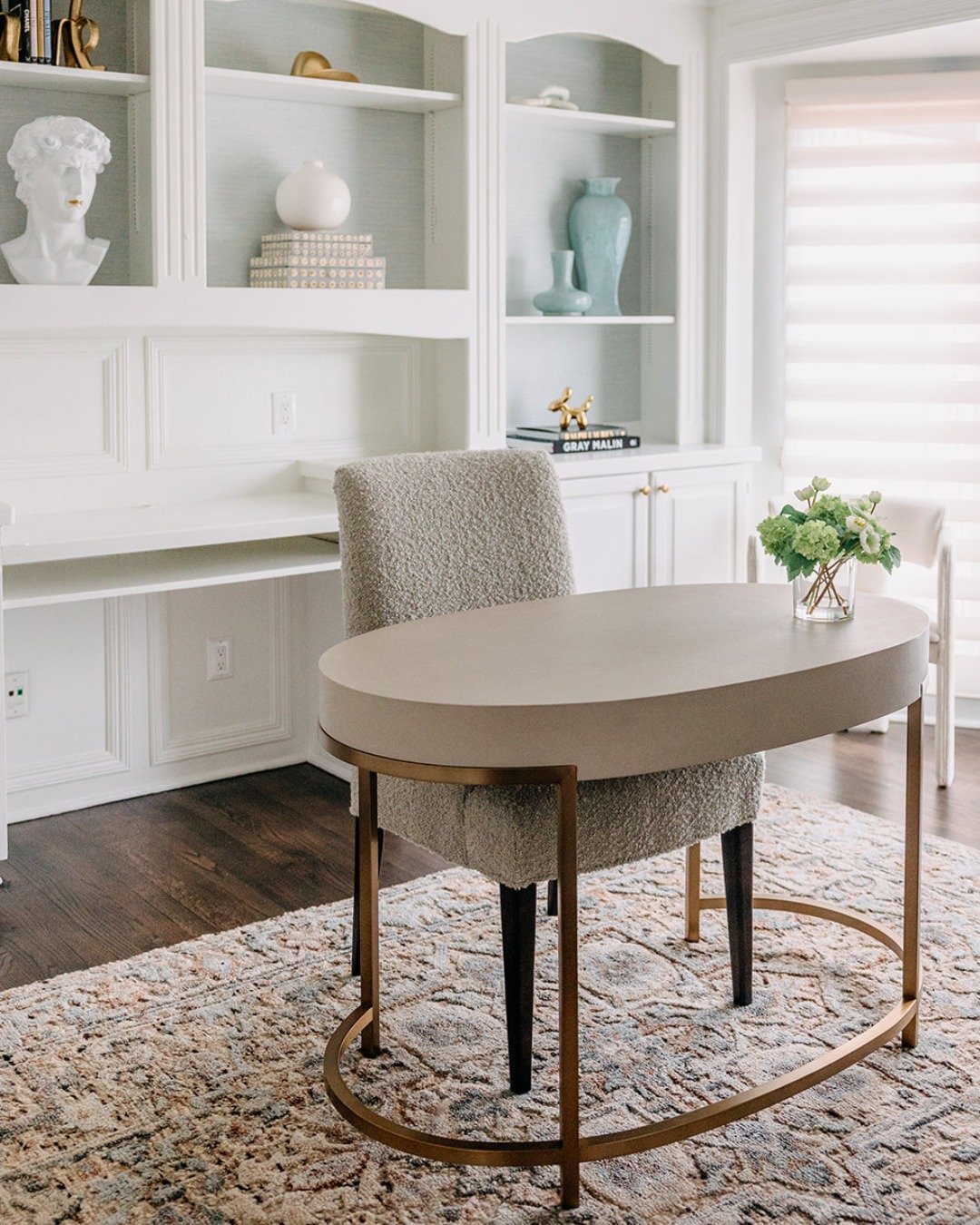 Bright white home office with built-in shelves, a sculptural oval desk with a brass base, and a furry beige boucle chair over a patterned rug.