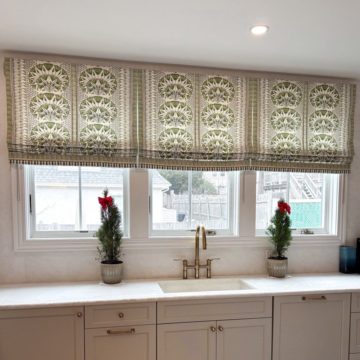 Kitchen sink area with white cabinets and a brass faucet, featuring a custom Roman shade in a repeating green and white geometric fan pattern acting as a decorative valance.