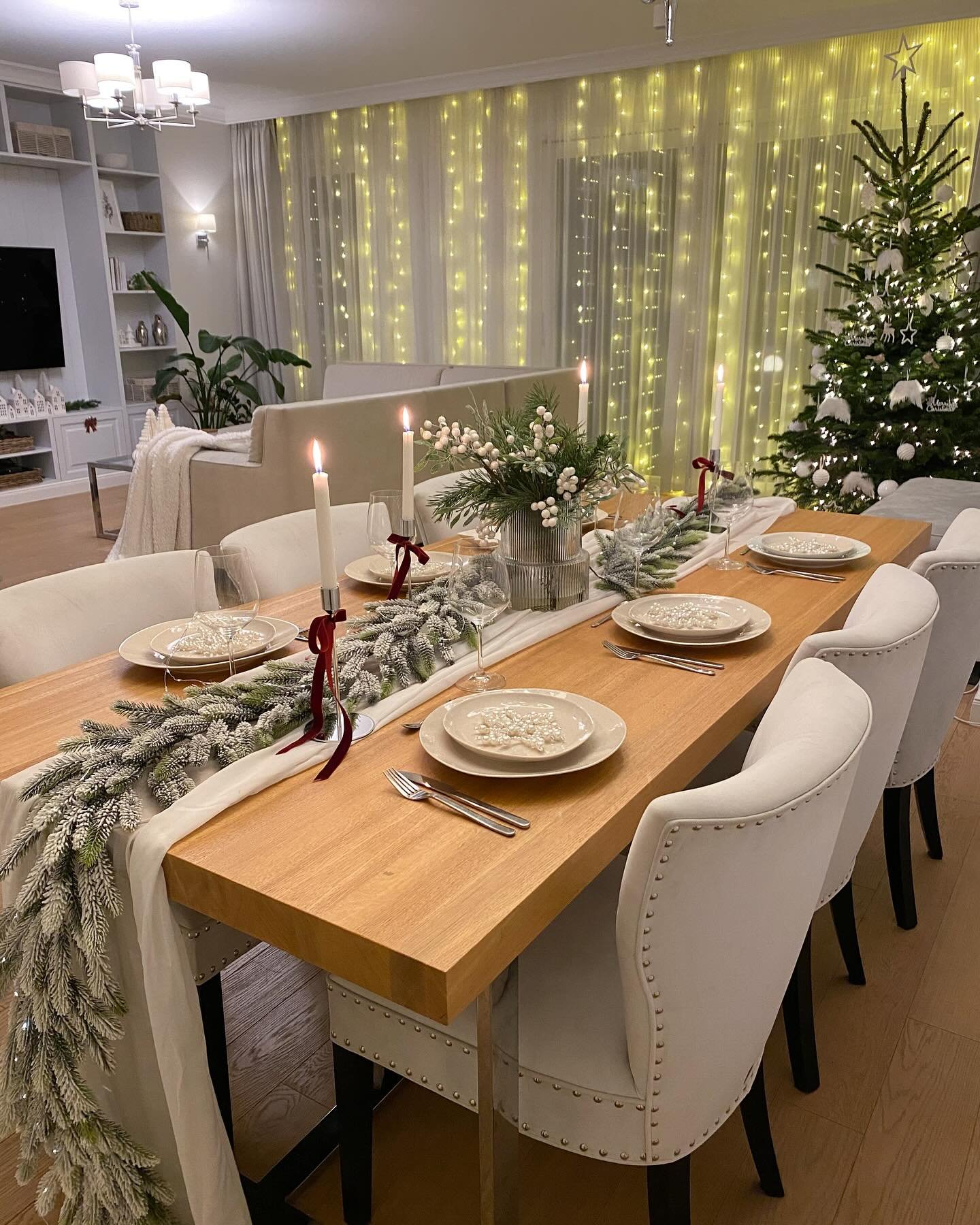 A modern dining table with a light wood surface and white upholstered chairs, set with a sheer white runner, snow-flocked evergreen garland, white beaded plates, and cream taper candles.