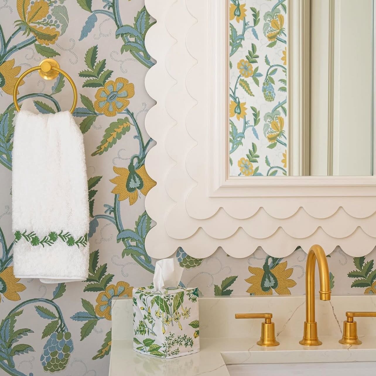 A polished gold towel ring holding a white hand towel with embroidered green trim against a patterned floral wallpaper.