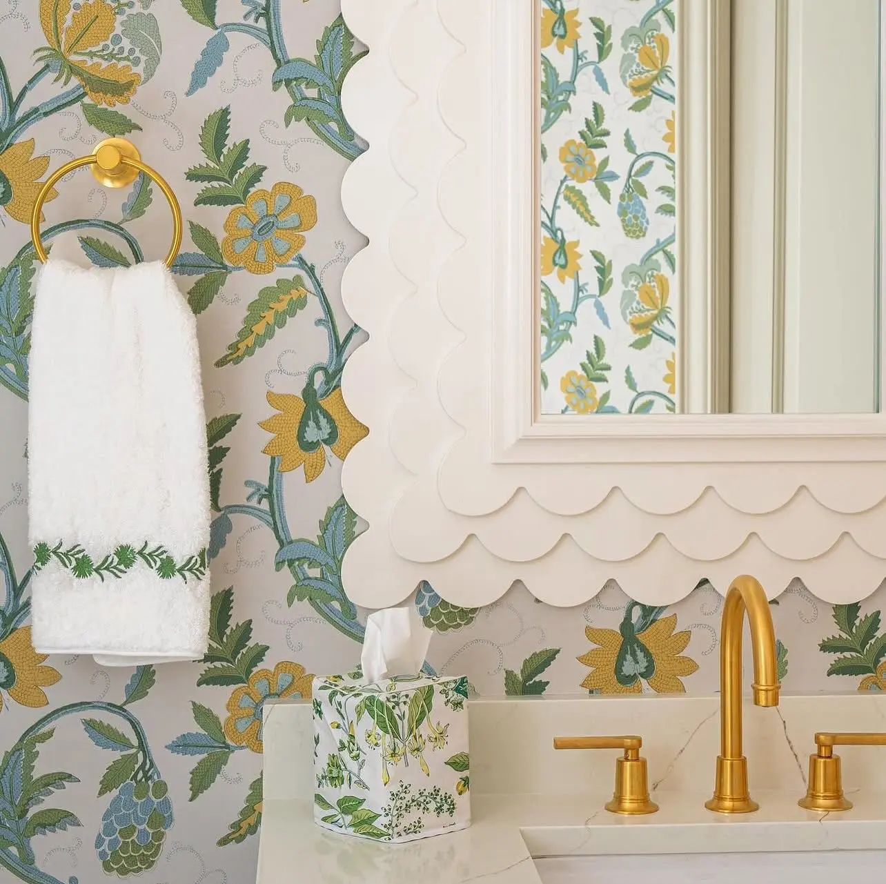 A polished gold towel ring holding a white hand towel with embroidered green trim against a patterned floral wallpaper.