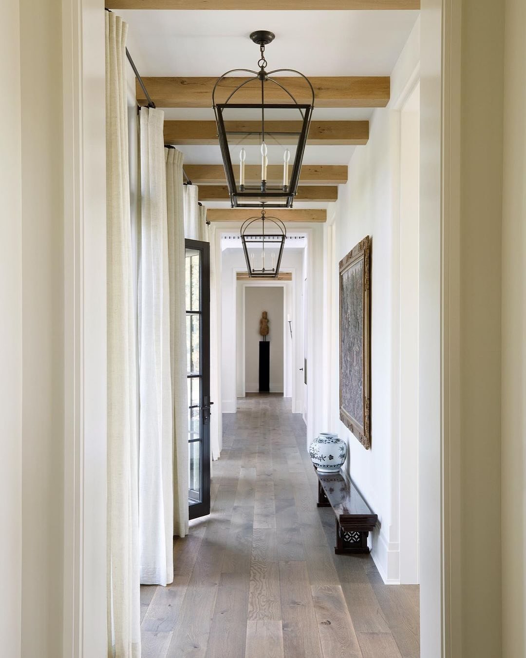 Long hallway with light wood floors, exposed natural wood ceiling beams, sheer drapes, and a series of large, black iron-framed lantern pendants.