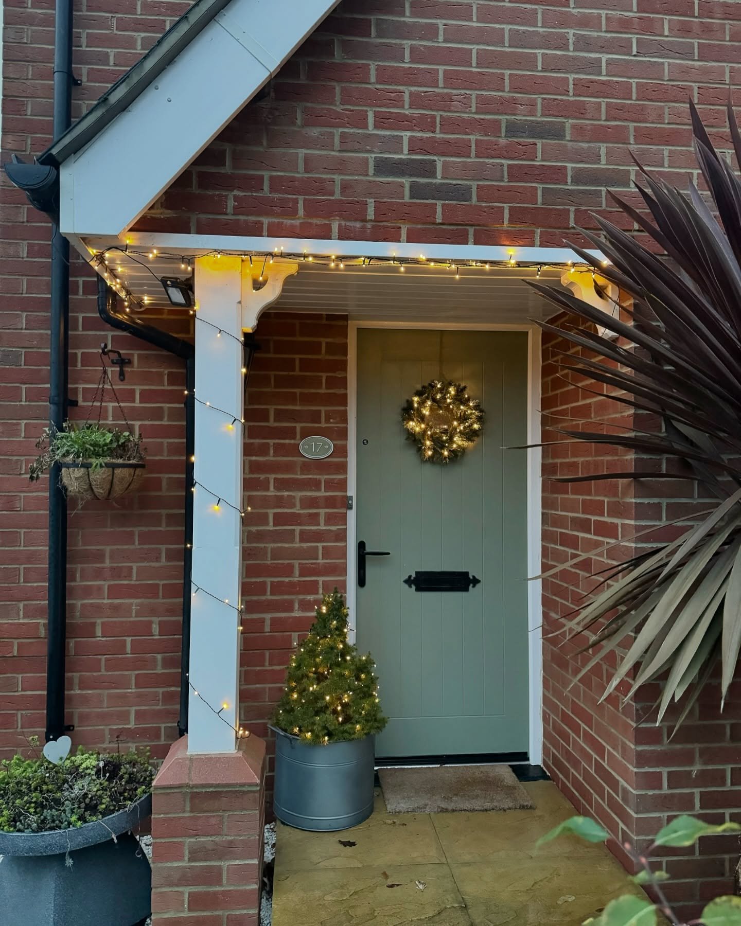 Warm white string lights wrap the white wooden column and roof of a small portico over a pale green front door with a lighted wreath and a small potted Christmas tree.