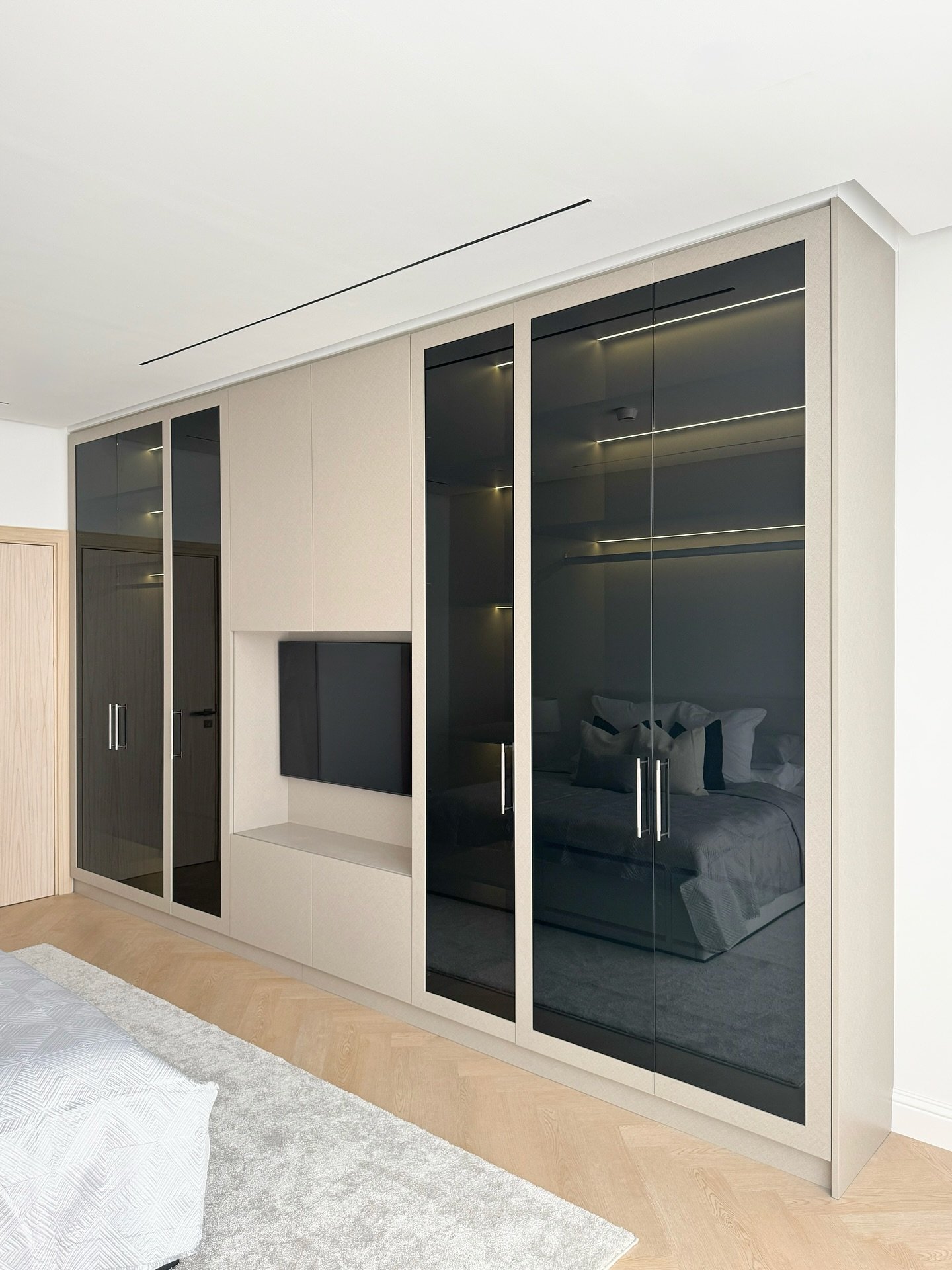 Modern full-wall wardrobe in taupe and black, featuring smoked glass doors and a central integrated media console for a television.