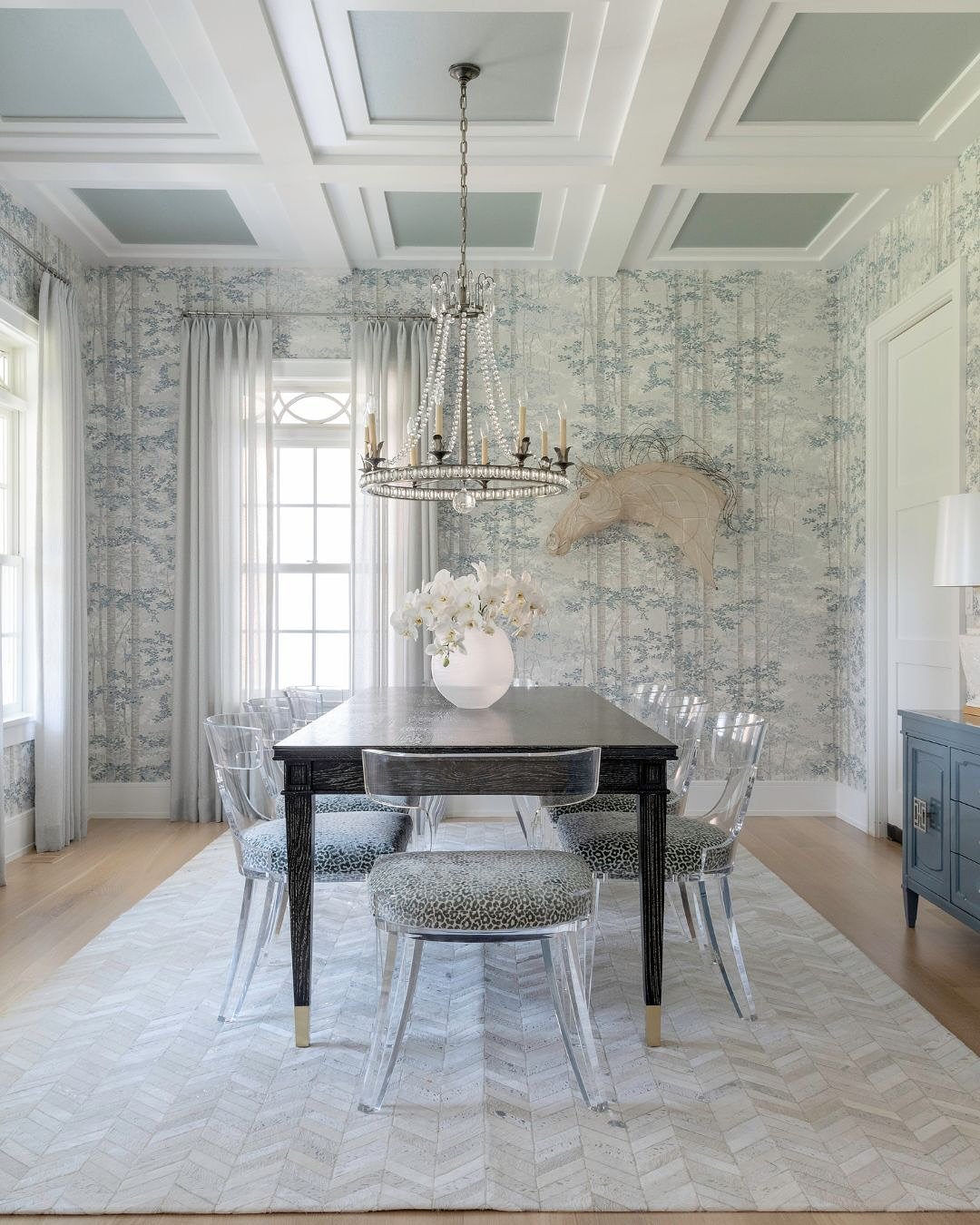 A formal dining room featuring light blue and white woodland-patterned wallpaper, a dark wood table, clear acrylic ghost chairs with patterned cushions, and a large beaded chandelier.
