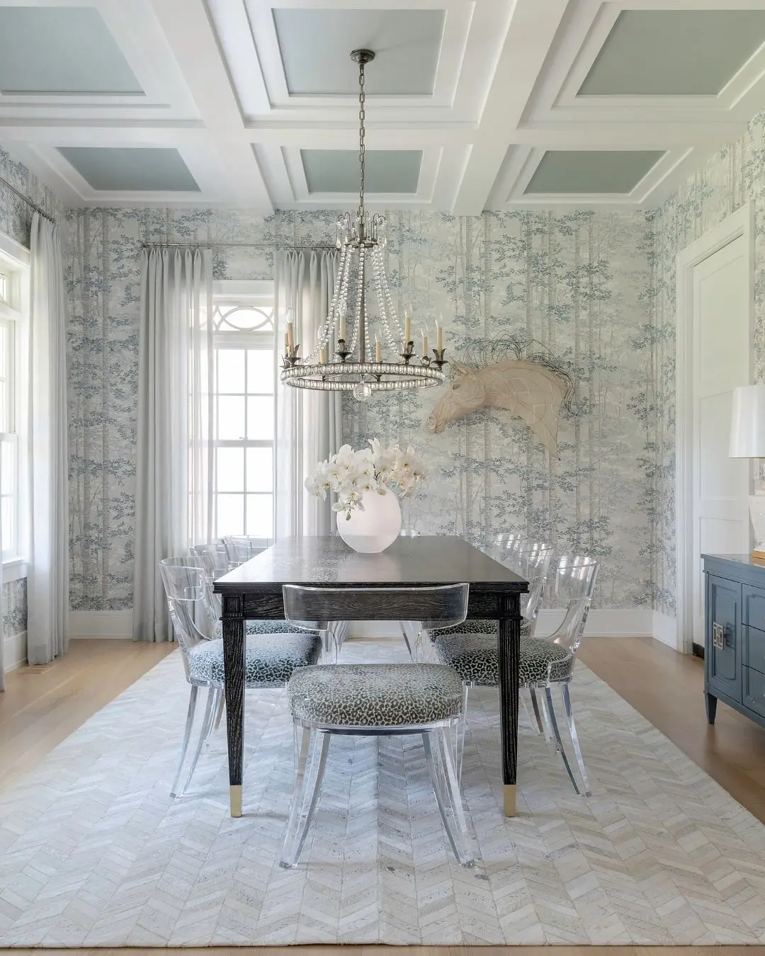 A formal dining room featuring light blue and white woodland-patterned wallpaper, a dark wood table, clear acrylic ghost chairs with patterned cushions, and a large beaded chandelier.