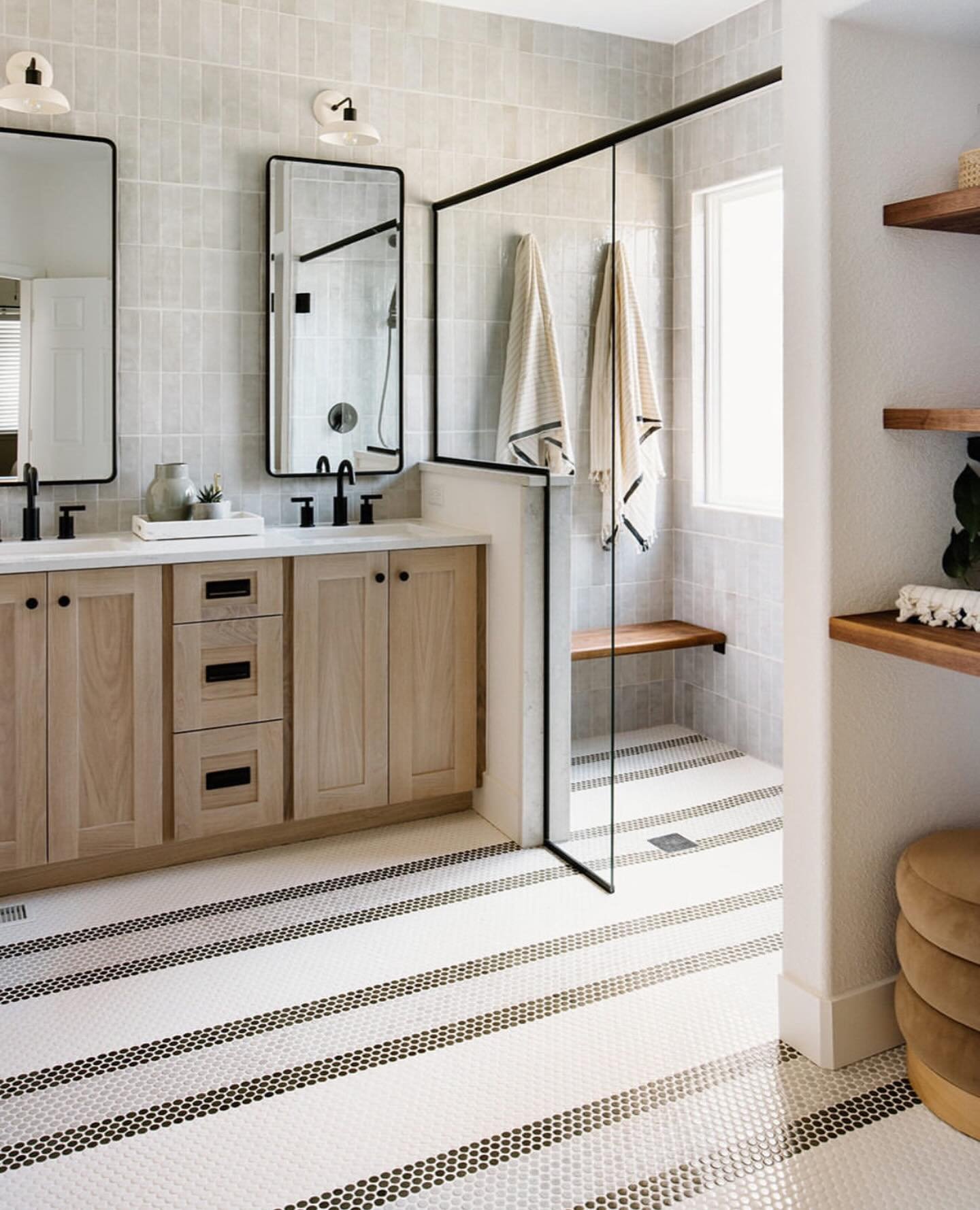 A modern bathroom with a light wood vanity and a white penny tile floor featuring parallel black-and-white striped accent lines running across the room.