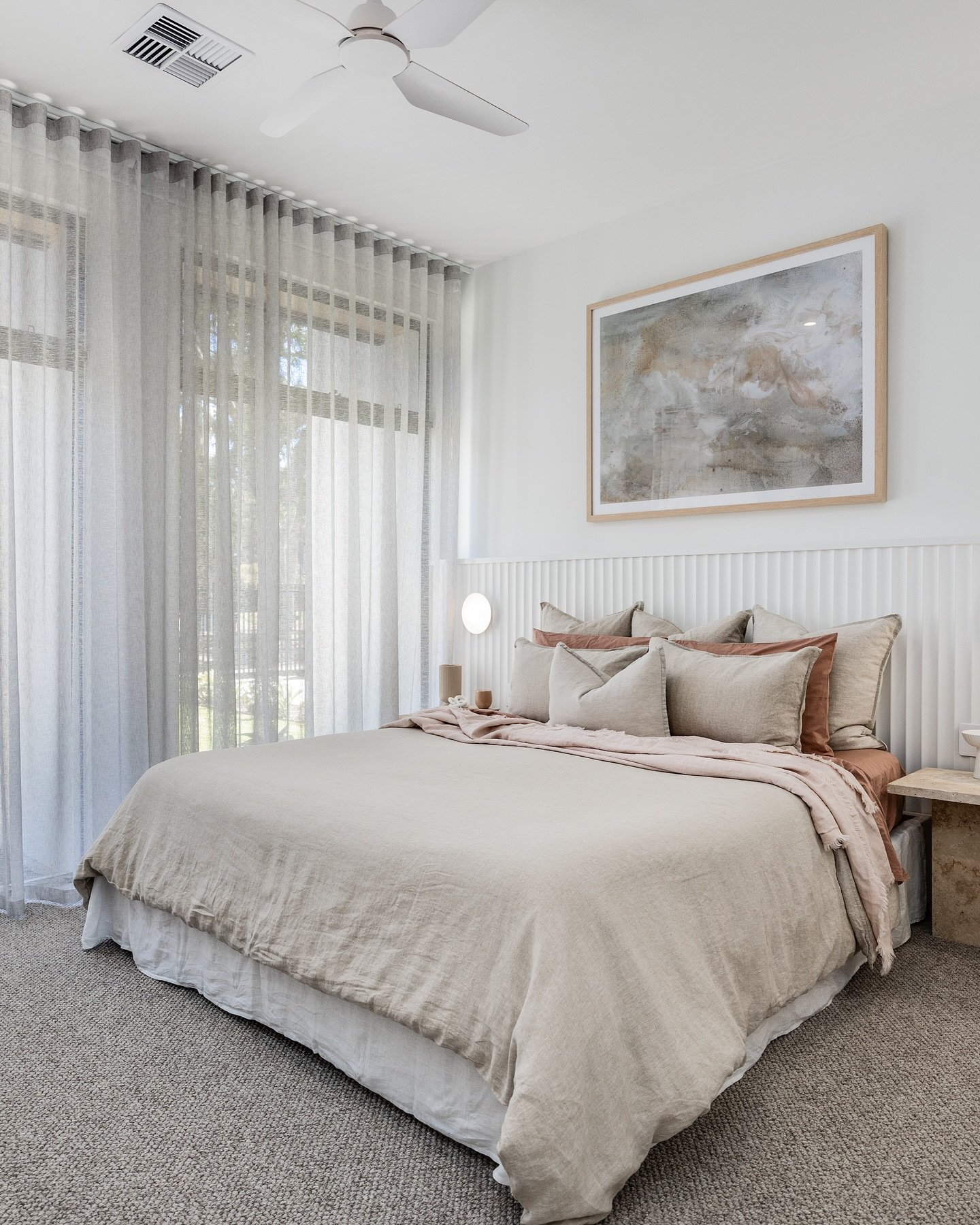 Bedroom featuring a fluted white wood panel accent wall behind the bed, layered linen bedding in shades of taupe and rust, and sheer grey curtains.