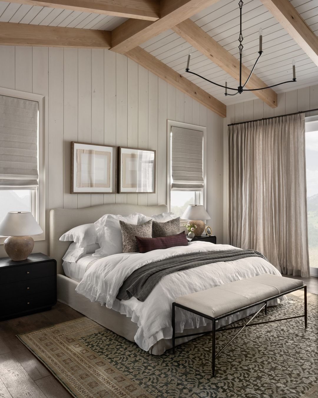 A luxurious bedroom with a vaulted ceiling and exposed light wood beams, shiplap walls, a large bed with white linens, dark nightstands, and a patterned area rug.