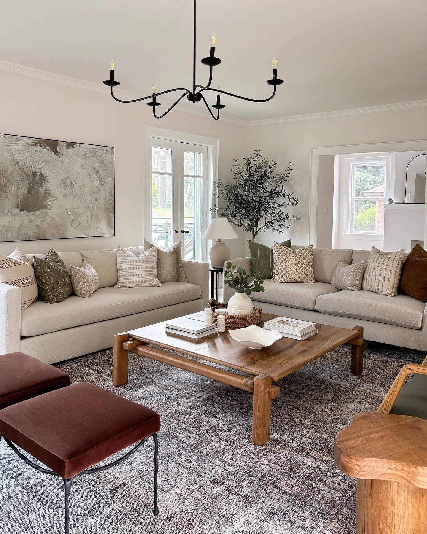 A neutral living room with two facing beige sofas, a large square wood coffee table, and a black minimalist chandelier hanging above.