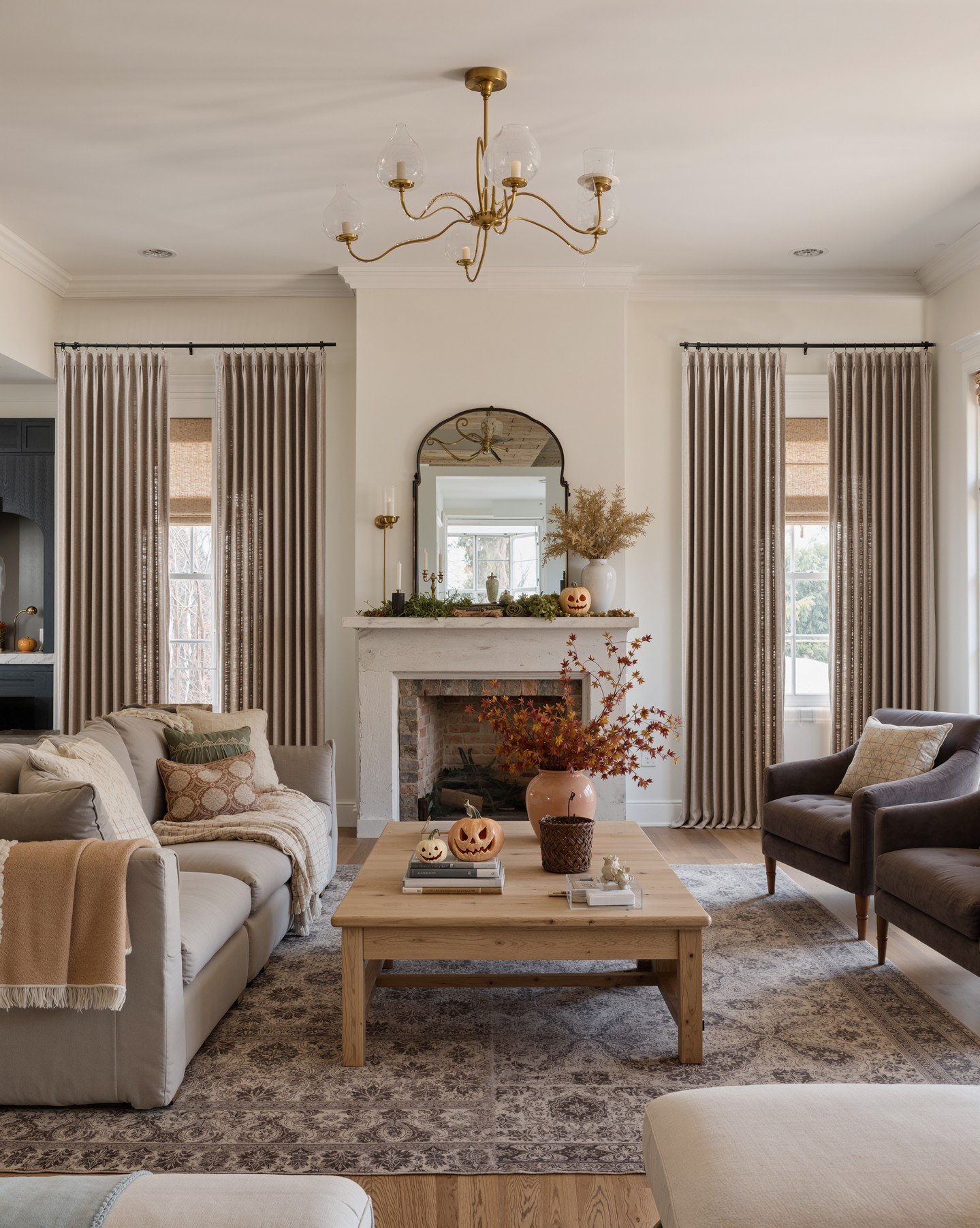 Two sets of floor-length, pleated taupe curtains framing a fireplace, layered over woven wood shades in a warm living room.