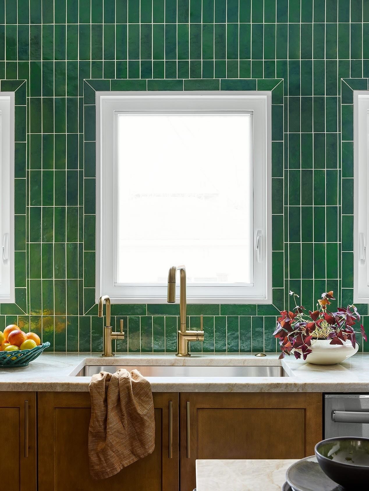 Kitchen backsplash featuring glossy, vertical dark emerald green subway tiles with light-colored grout surrounding a window, paired with brown wood cabinets.