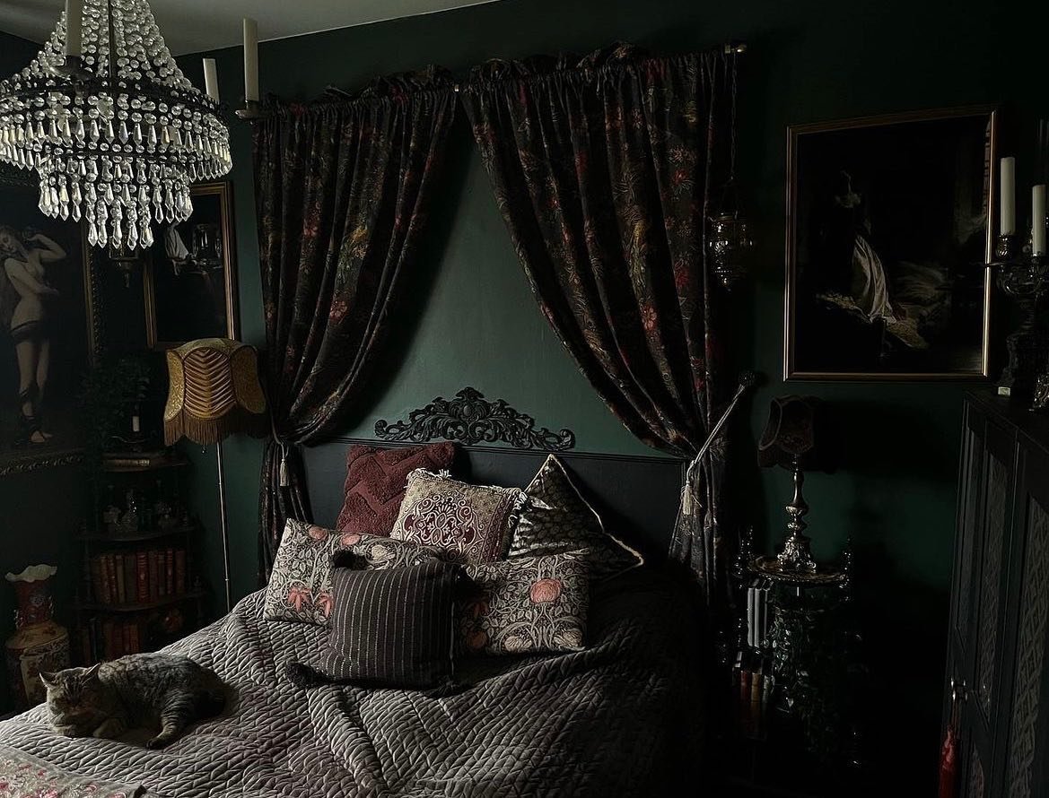 Dark, maximalist bedroom with deep emerald green walls, heavy floral velvet curtains, a dark carved headboard, and an ornate crystal chandelier.