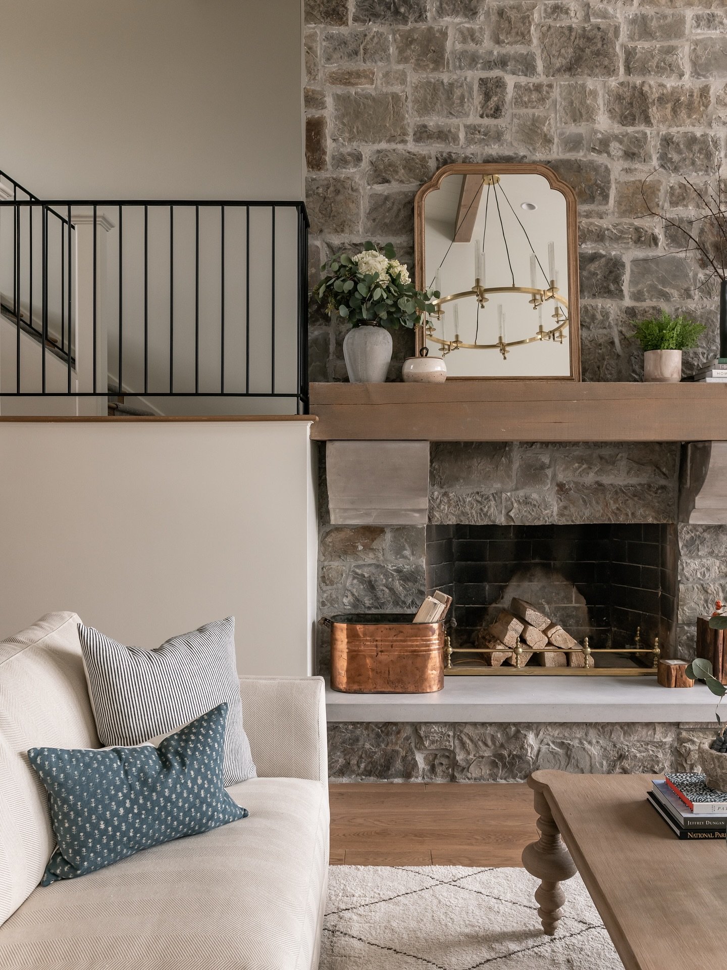 A full-height, floor-to-ceiling fireplace constructed from rough-cut grey fieldstone with a thick, natural wood mantelpiece and a copper log holder on the hearth.
