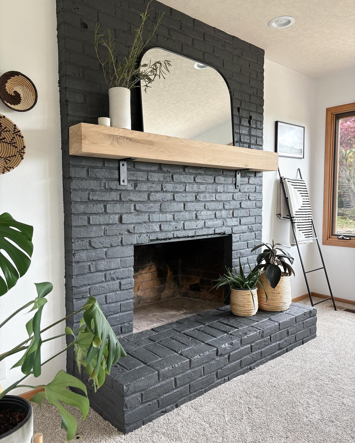 A painted charcoal gray brick fireplace with a light wood floating mantel. A black arched mirror and potted plants decorate the mantel and hearth.