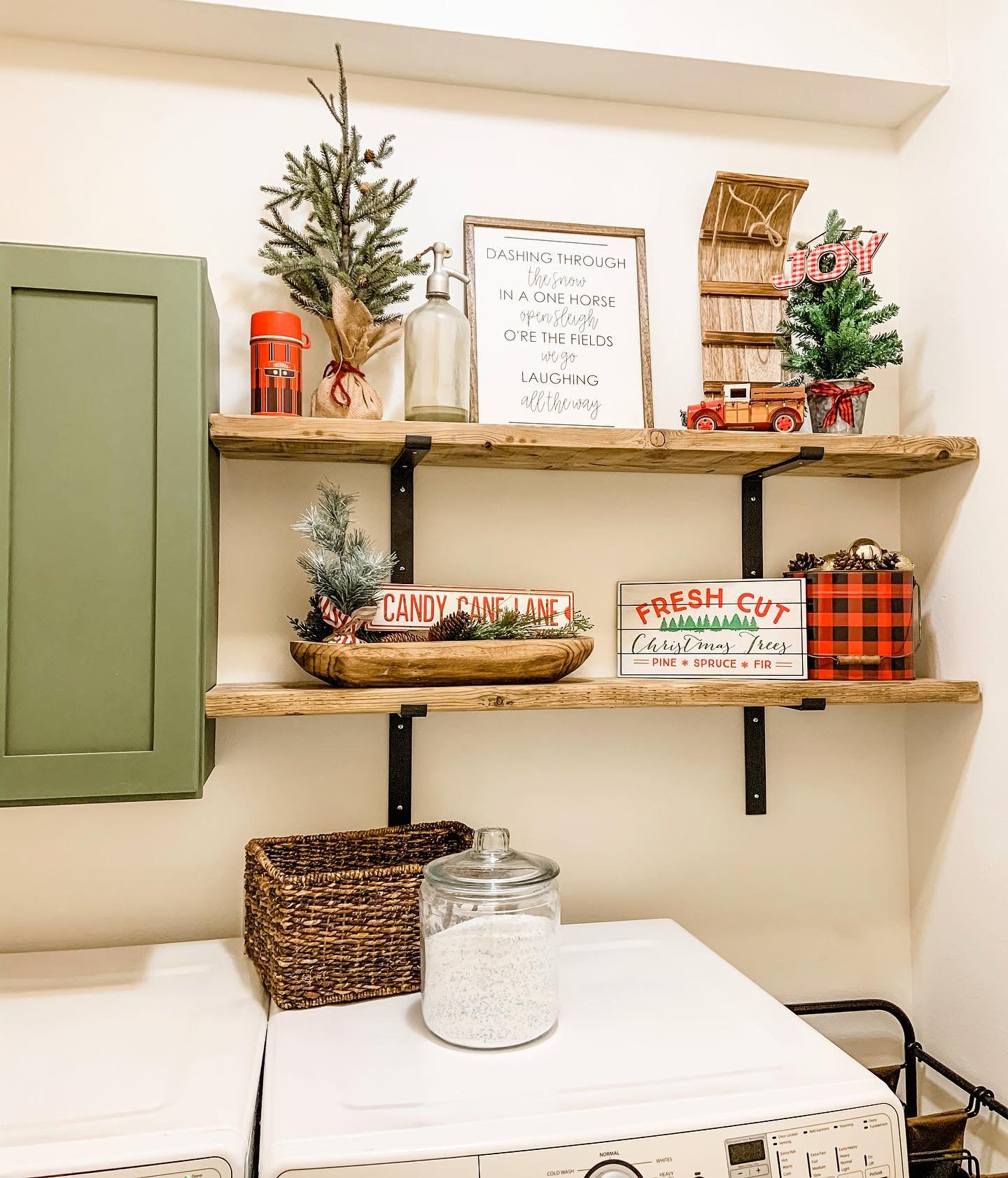 Wooden laundry shelves decorated with small pine trees and holiday signs above a washing machine