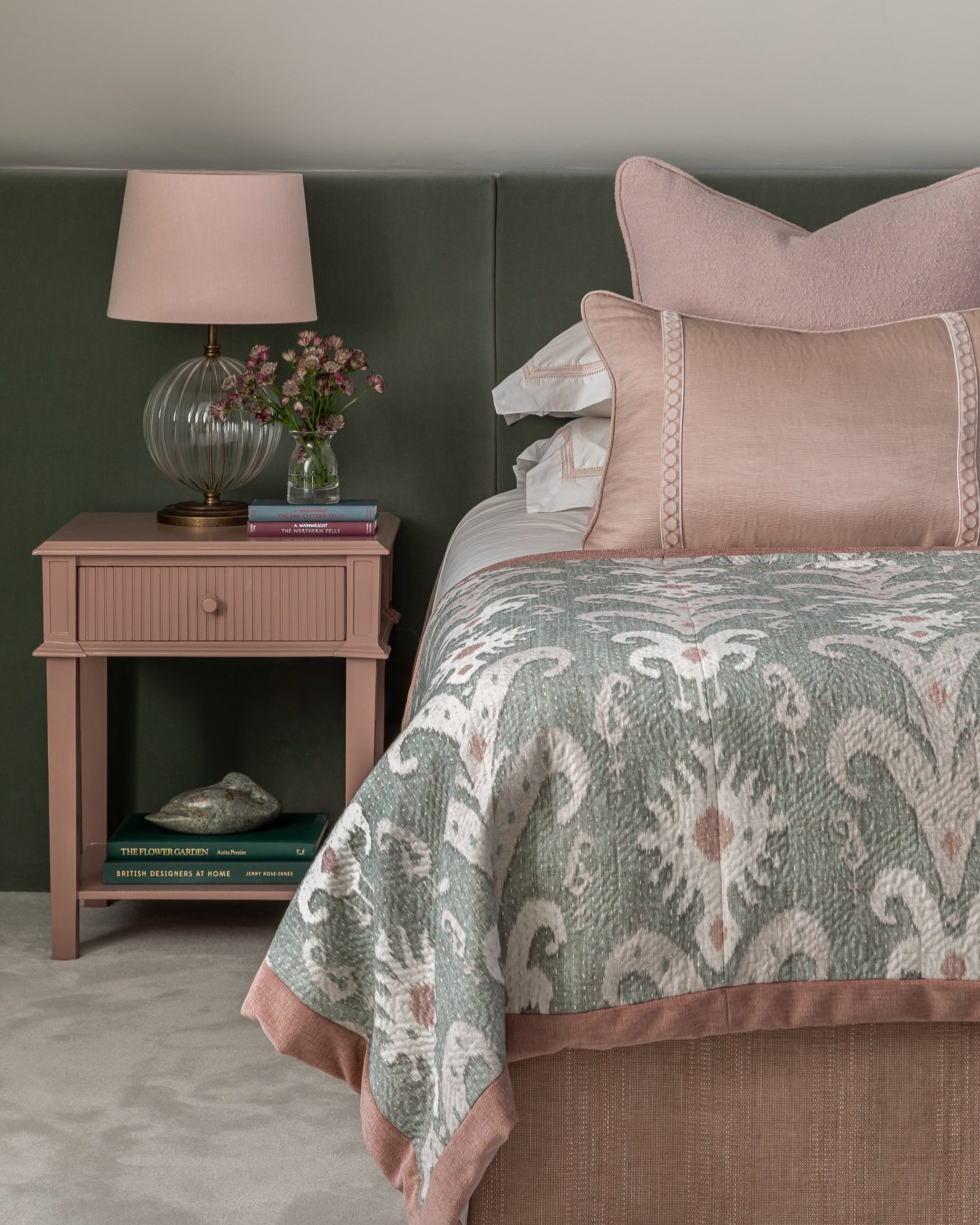 A cozy bedroom featuring a sage green upholstered headboard, a muted pink nightstand with a matching lamp, and a patterned quilt.