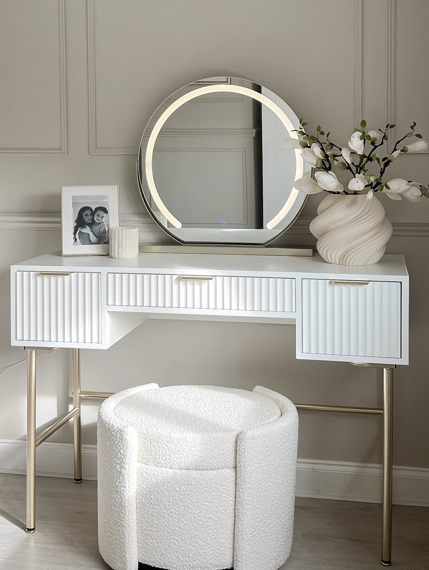 A white dressing table with fluted drawers and gold legs, paired with a round LED-lit mirror and a textured white bouclé stool, against a panelled wall.