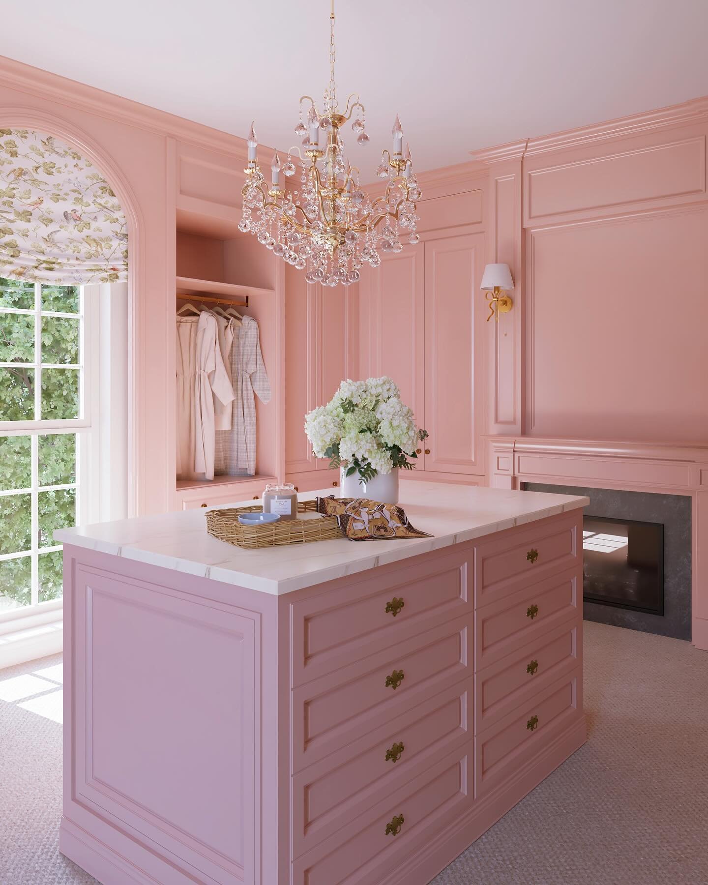 A luxurious walk-in closet featuring light pink walls, a large pink central island with a marble top, and a gold crystal chandelier.