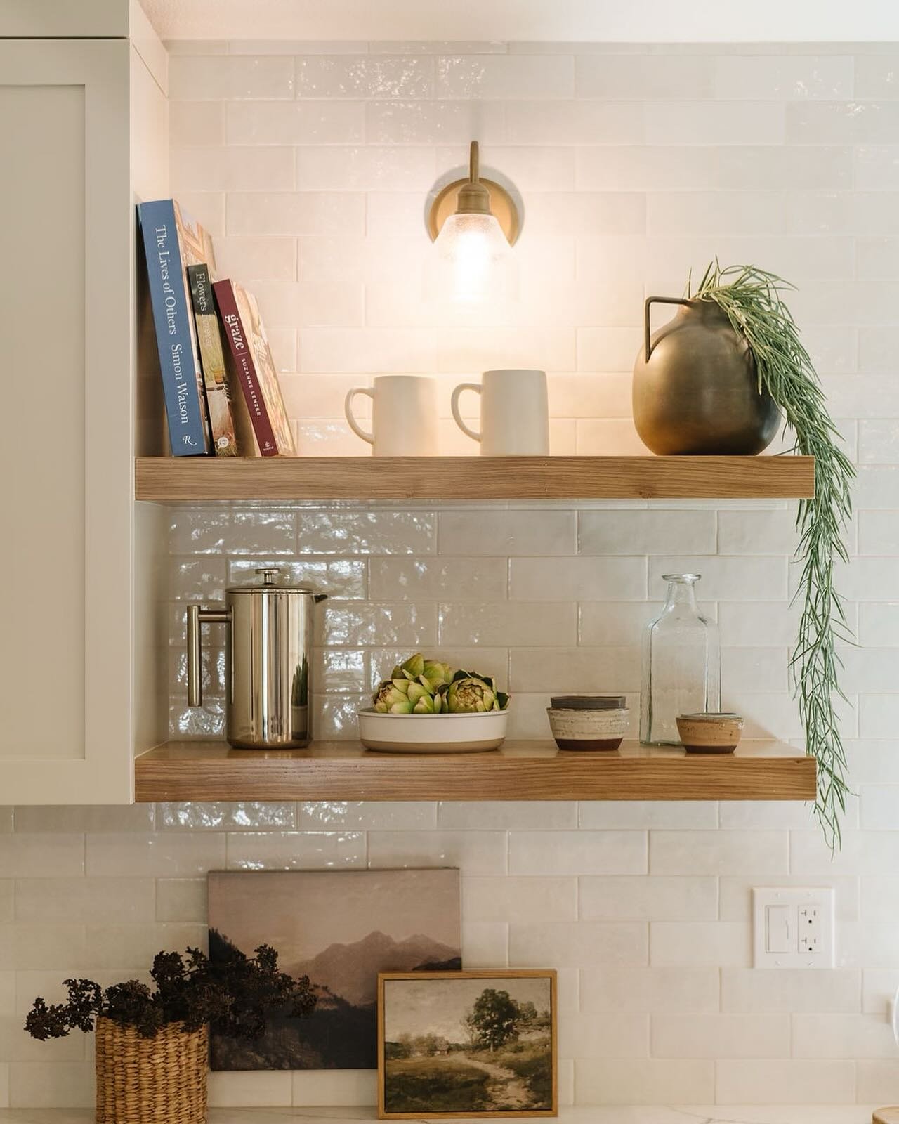 Light wood floating shelves in a kitchen vignette with a brass sconce, cookbooks, and decor.