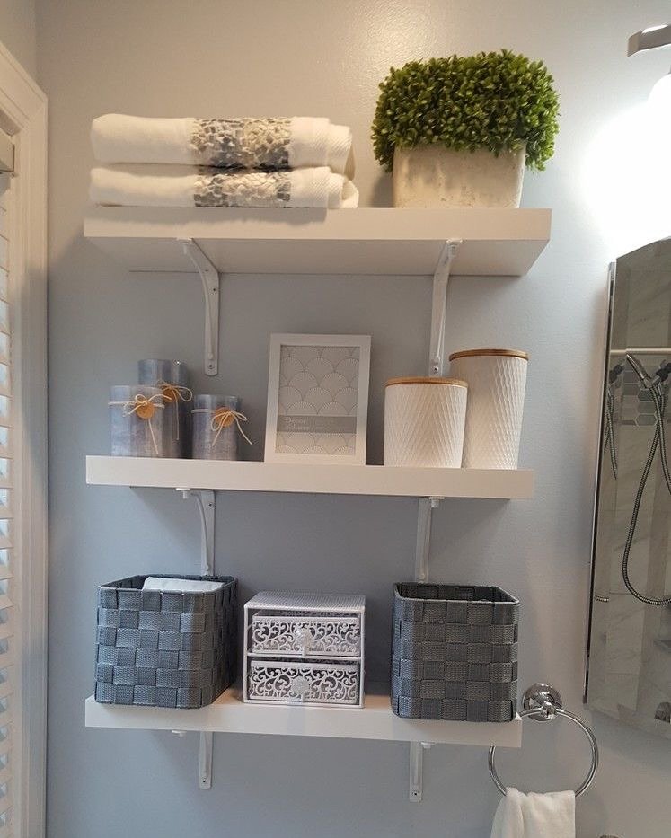 Three white shelves mounted with visible white brackets on a blue wall, holding baskets, towels, and canisters.