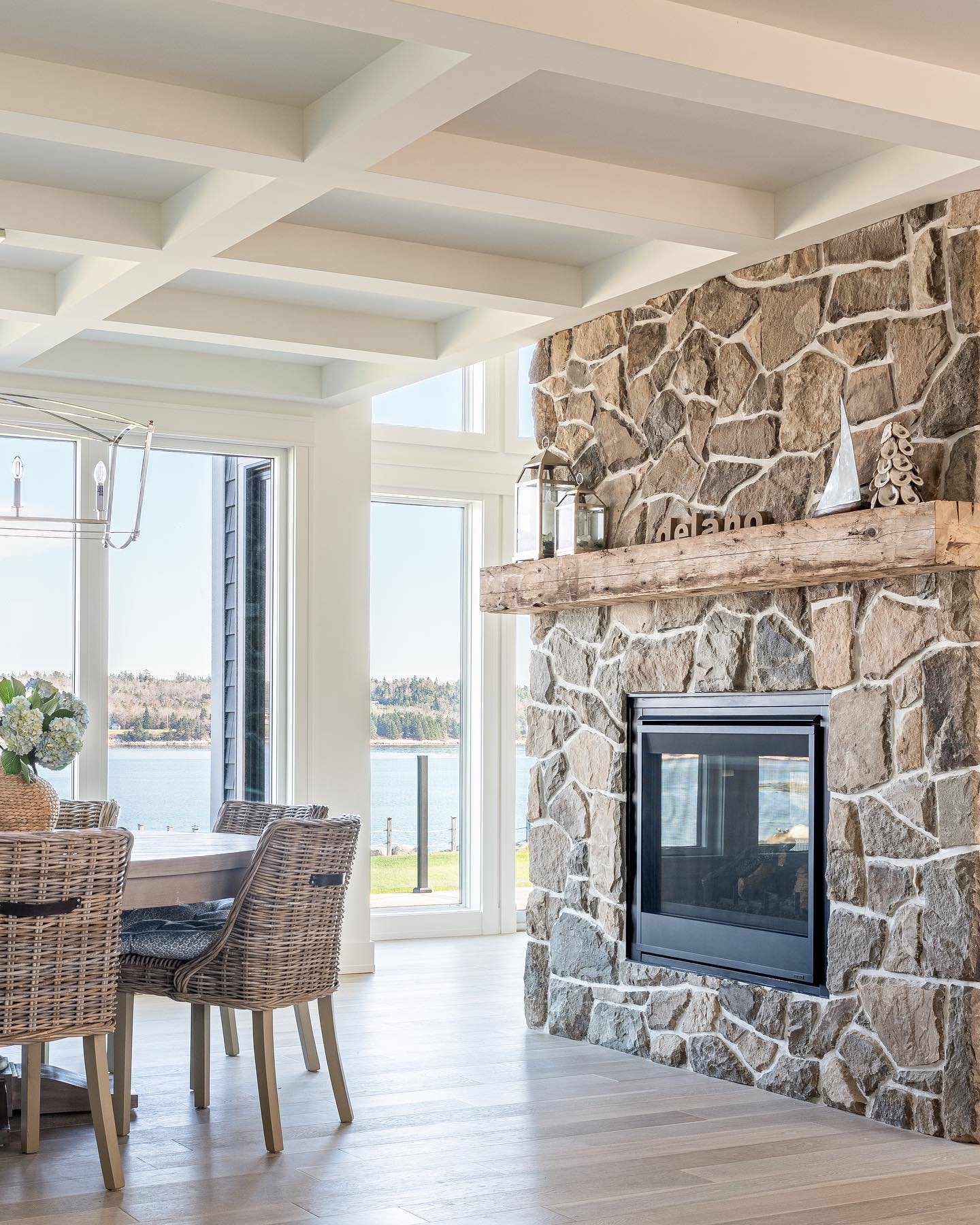A full-height, rustic fieldstone fireplace in a dining room, featuring a thick, natural-edge wood mantel, a black gas firebox, and large windows offering a clear view of a lake or ocean.