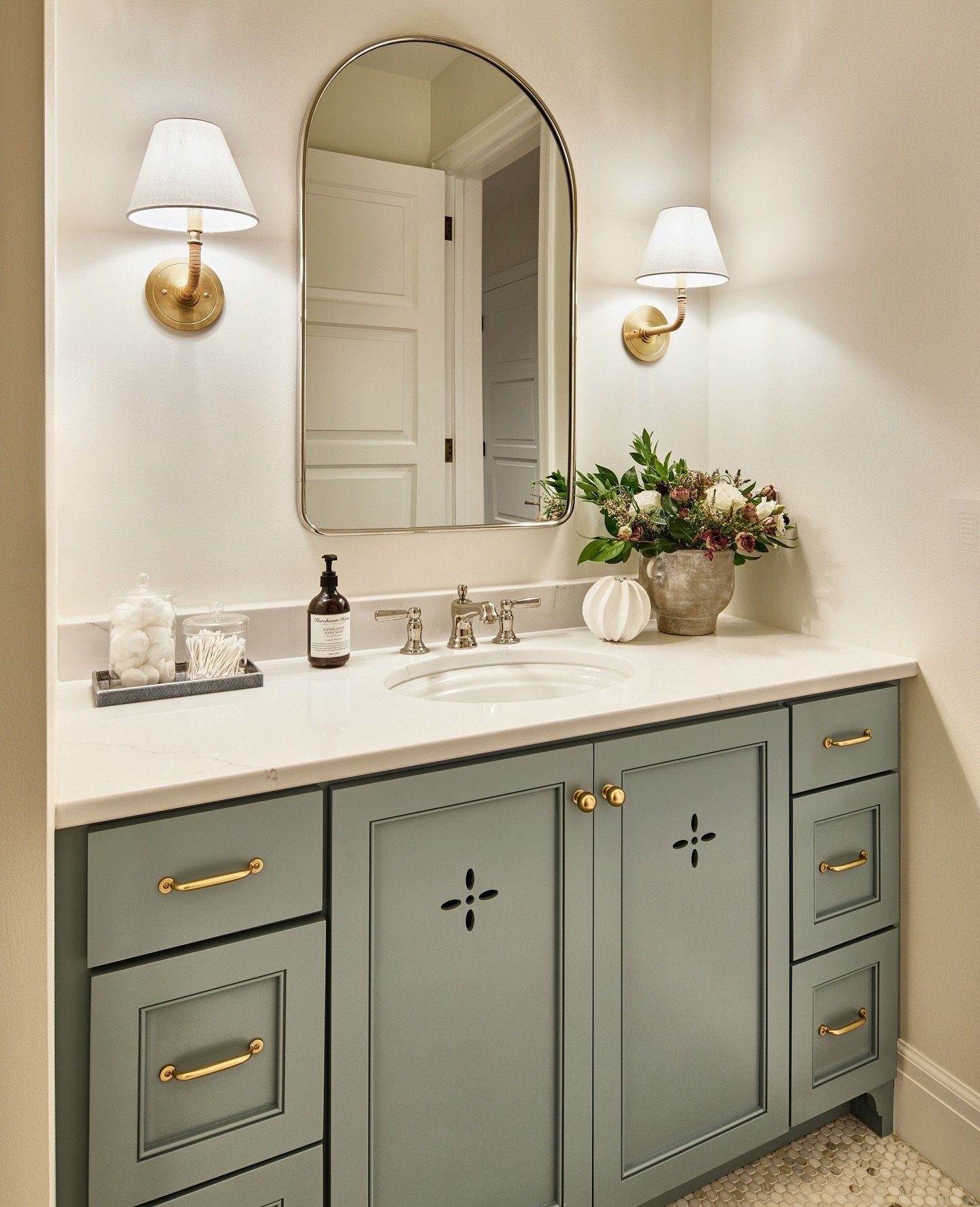 White fabric shade sconces with curved brass arms flanking an arched mirror above a pale blue-green vanity with gold hardware.