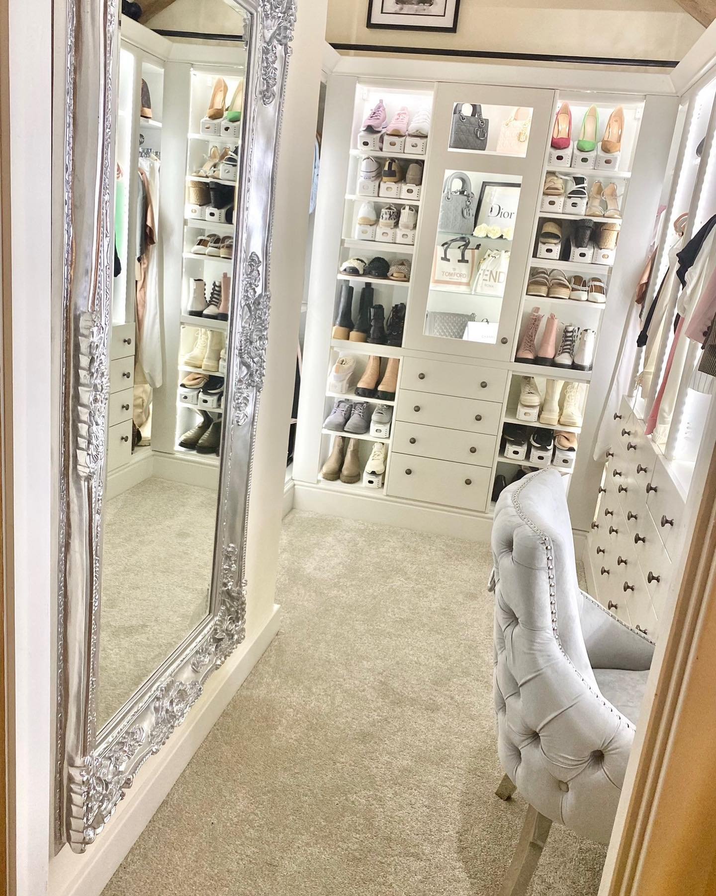 A cozy, neutral-toned walk-in closet focused on shoe storage, featuring a large, ornate silver mirror and a tufted grey chair.