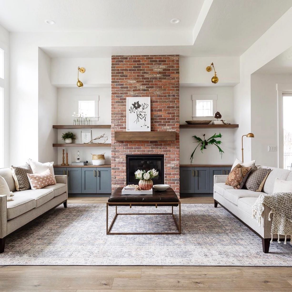 A living room featuring a floor-to-ceiling brick fireplace with natural red and brown tones. It is flanked by blue-gray built-in cabinets and floating wood shelves.