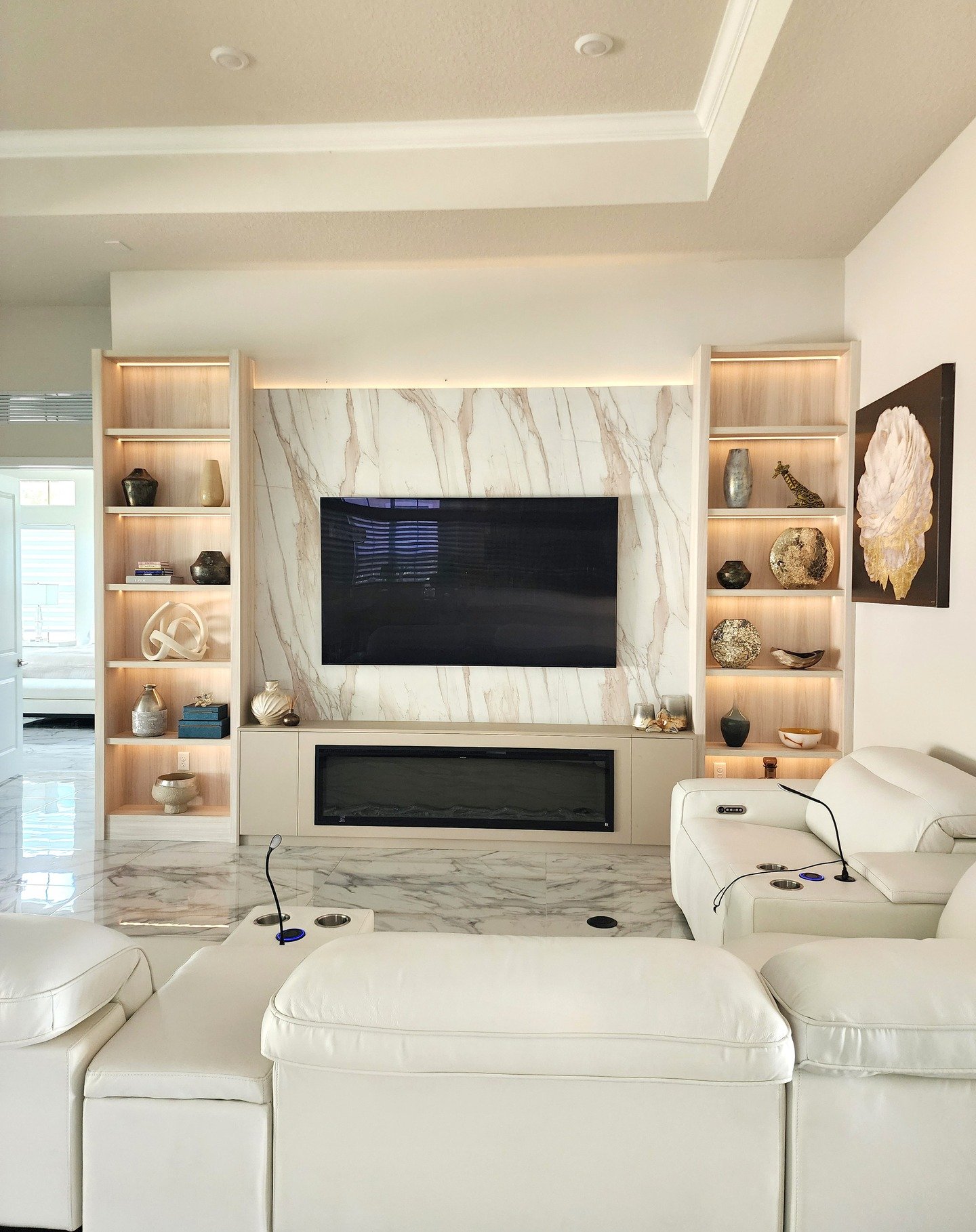 Tall light wood shelves with LED lighting flanking a marble TV wall with an electric fireplace.