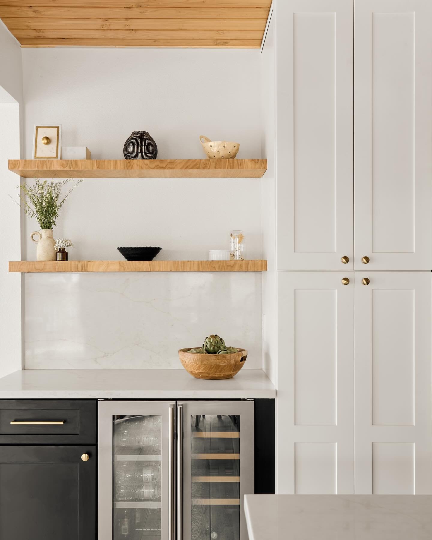 Two light wood floating shelves mounted on a white marble kitchen backsplash next to white pantry cabinets.