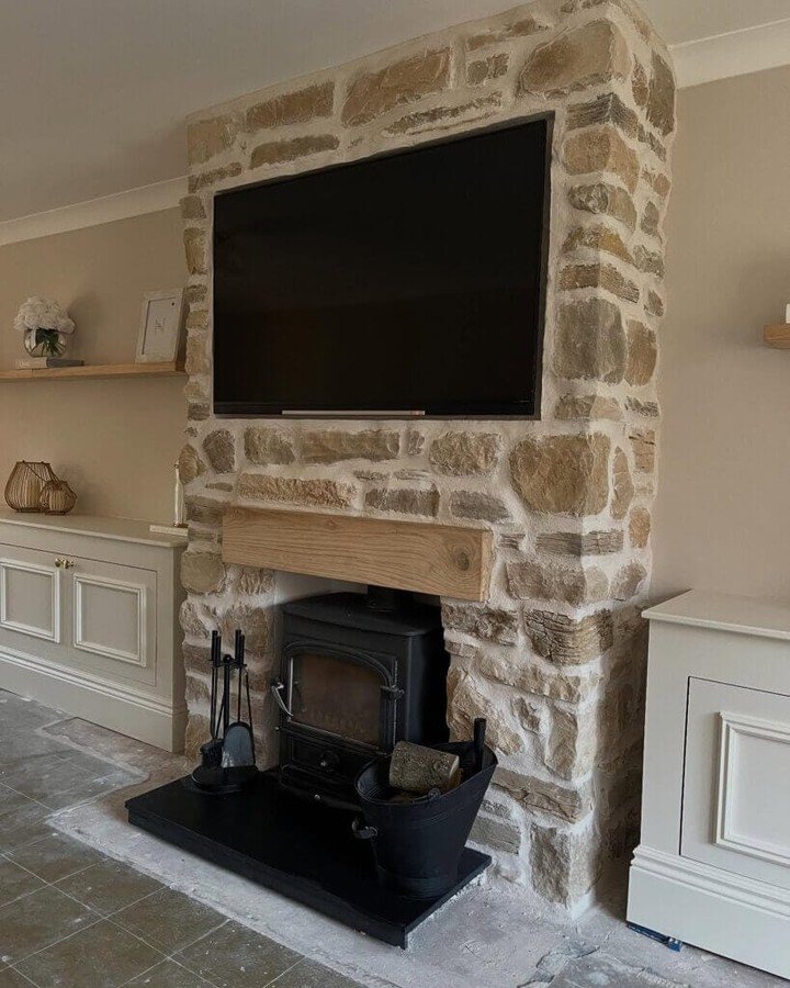 A chimney breast made of chunky, pale tan and cream rustic stone, featuring a small wooden mantel shelf, a black wood-burning stove on a dark hearth, and a flat-screen TV mounted above.