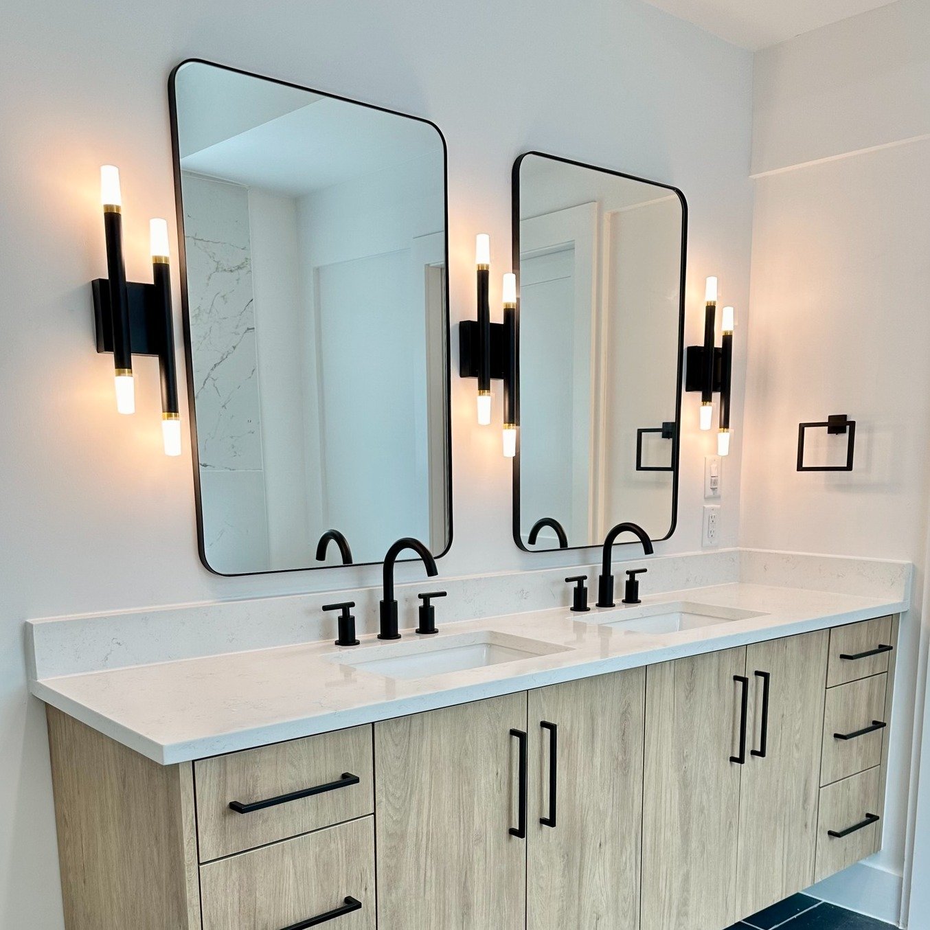 Vertical black and gold wall sconces with multiple light bars flanking two rectangular black-framed mirrors over a light wood double vanity.