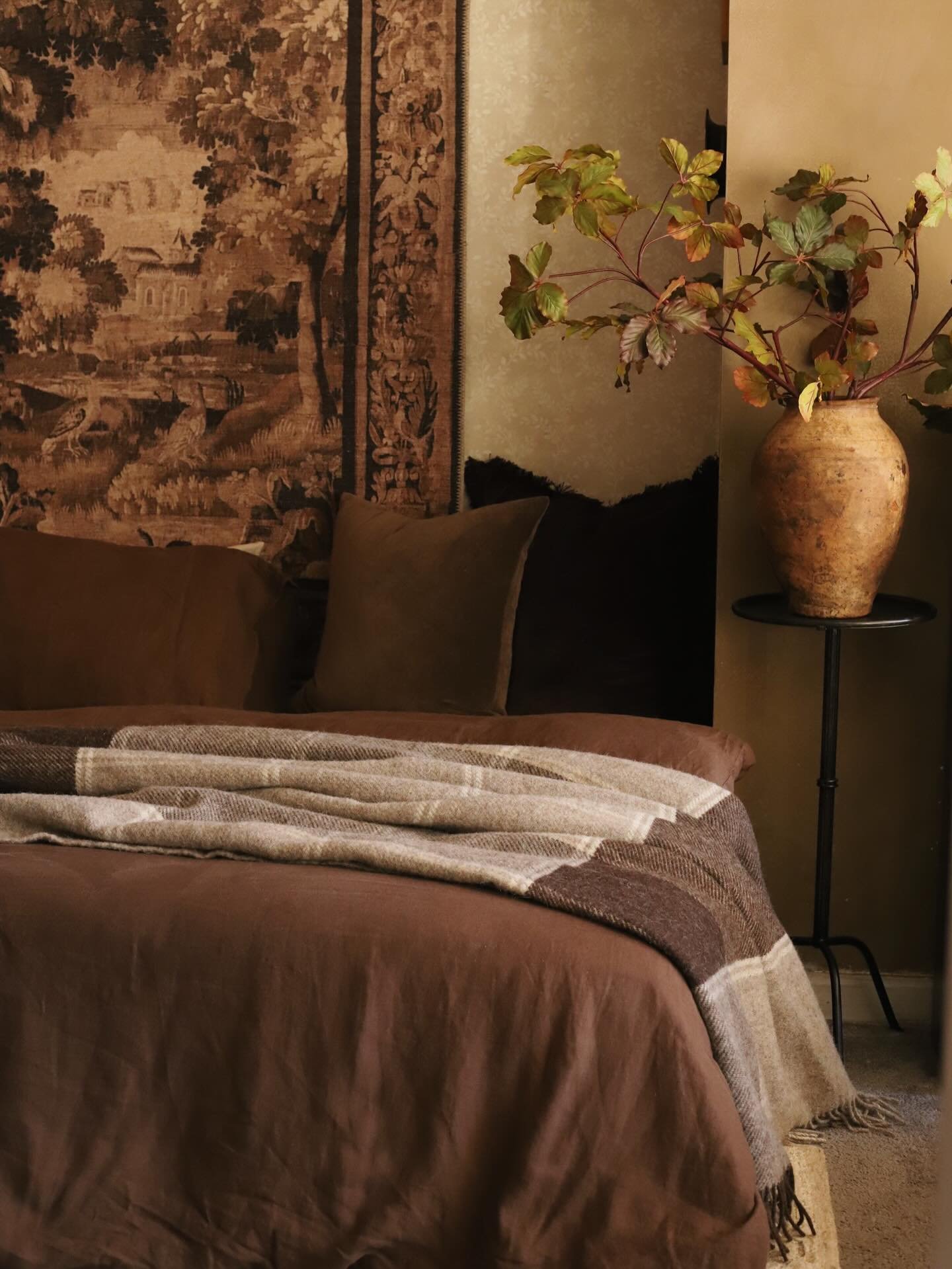 Moody bedroom featuring a large antique wall tapestry, rustic clay vase with branches, and dark brown bedding
