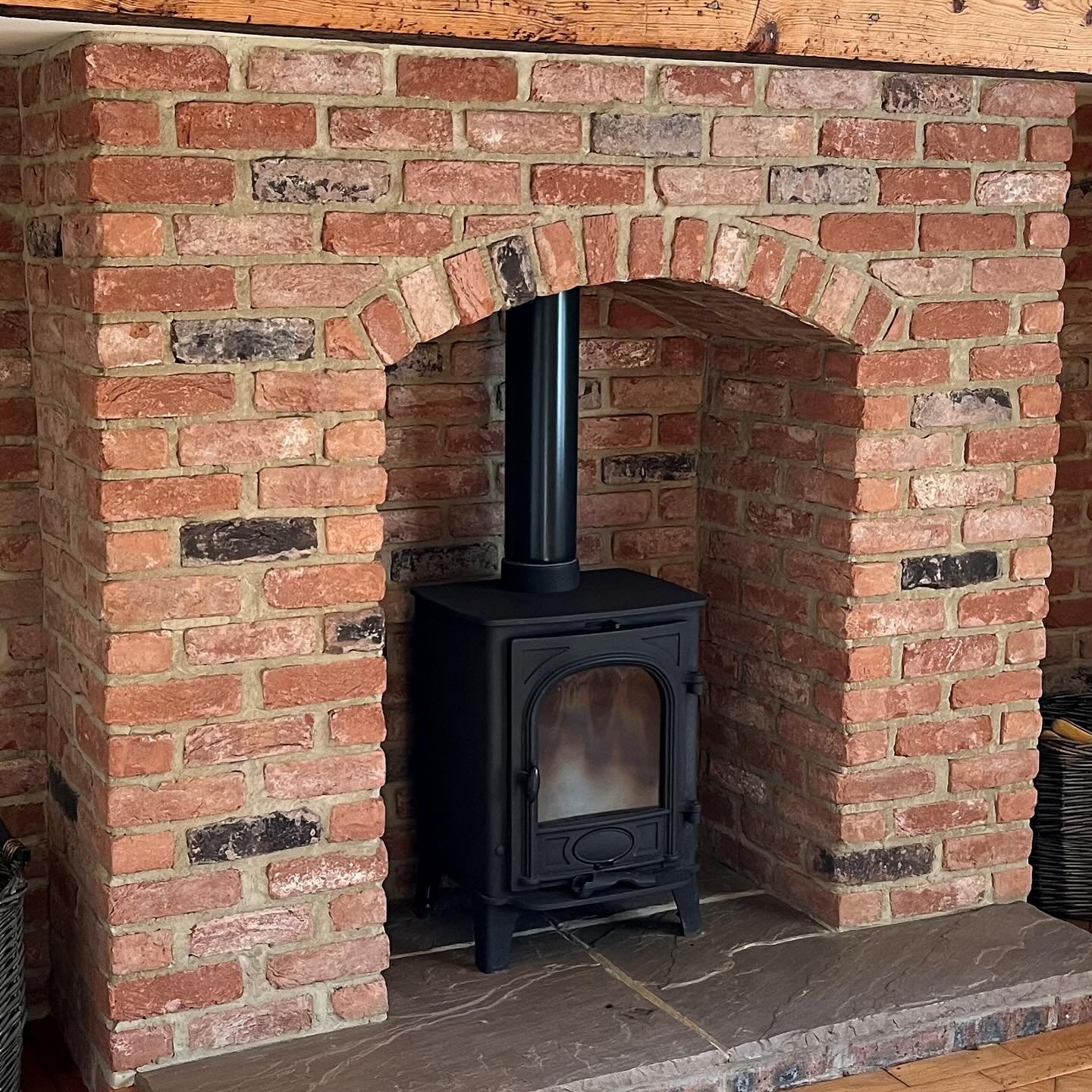 A close-up of a rustic brick fireplace with an arched opening, housing a black freestanding wood-burning stove on a dark stone hearth.