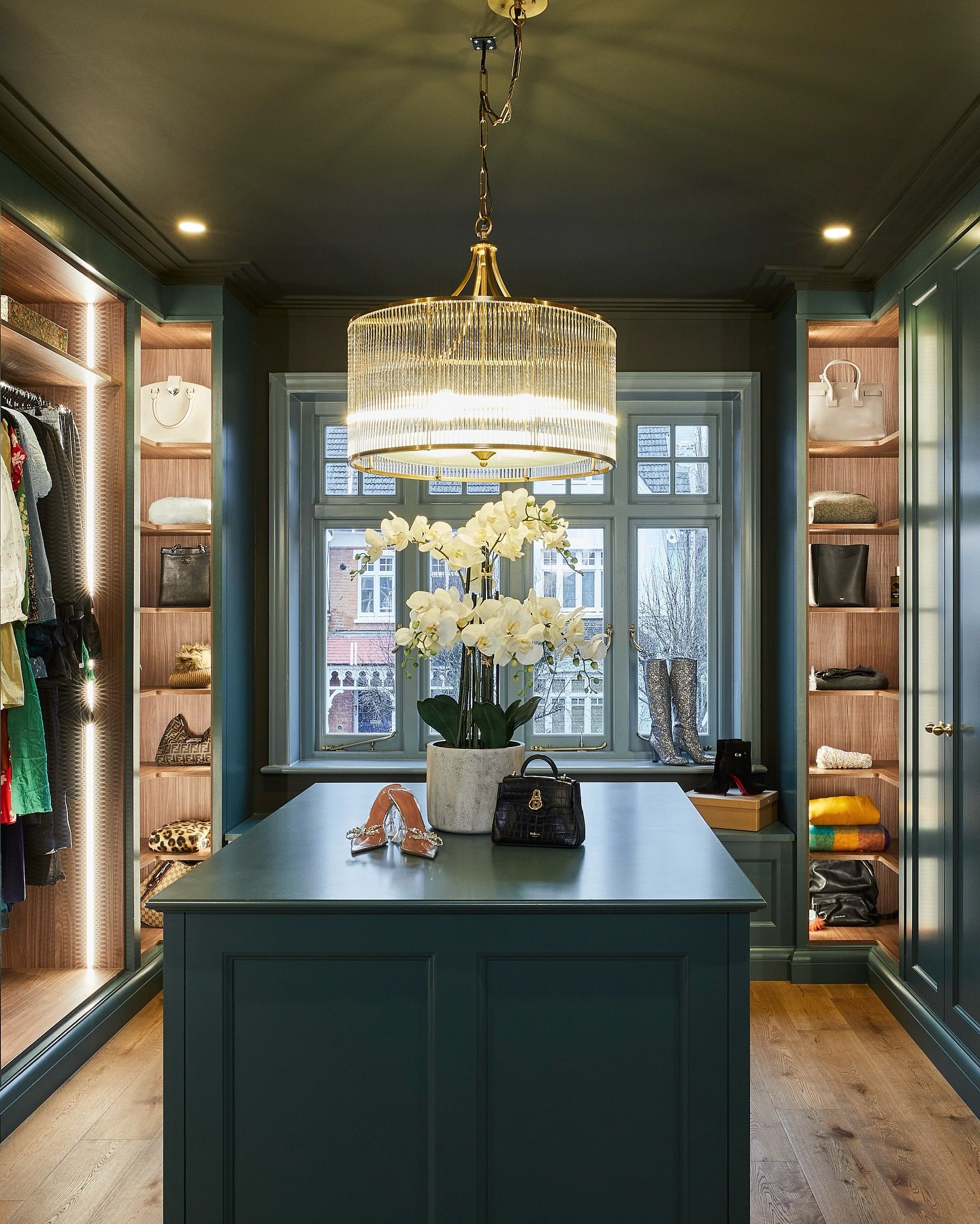 A luxury walk-in closet featuring cabinetry and walls painted in a deep forest green, a matching central island, and a gold and glass rod chandelier.